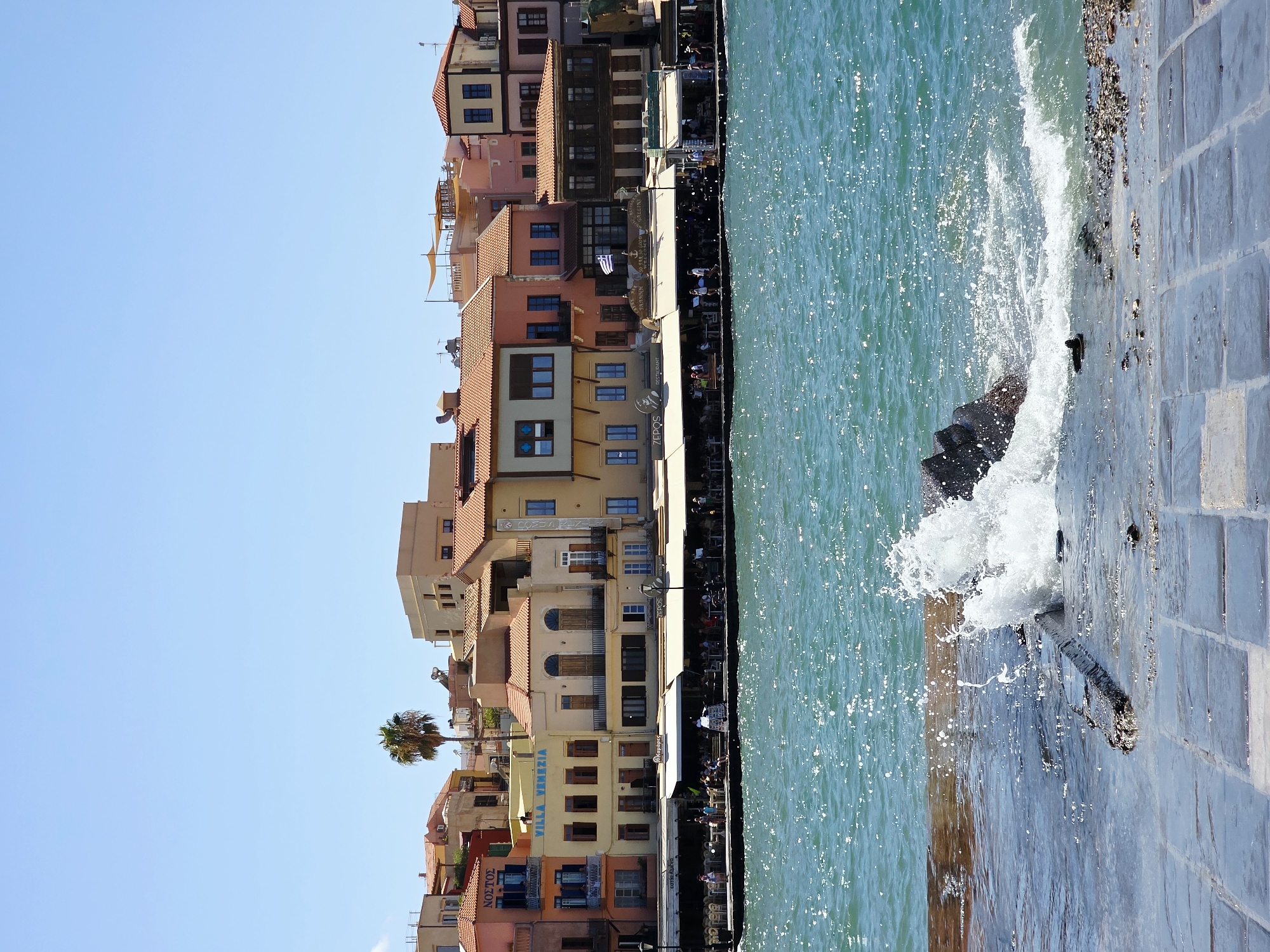 The image captures a sunny day scene of the Venetian harbor in Chania, Greece. In the foreground, a wave crashes against the stone quay, creating a splash of white water. The quay is paved with large, rectangular stones, some of which are wet and reflective. Beyond the quay, the turquoise waters of the harbor are alive with sparkling sunlight.
Along the waterfront, a row of colorful buildings stands, representing the iconic Venetian architecture of Chania. These buildings are painted in various hues of terracotta, yellow, pink, and beige, with red-tiled roofs. Many of them feature balconies and arched windows.
Several establishments, likely restaurants or cafes, are set up along the waterfront, with awnings providing shade. People are visible sitting at tables and strolling along the promenade in front of the buildings. Signs in Greek and other languages are displayed, advertising businesses such as "NOSTOS" and "VILLA VENEZIA". A single palm tree stands tall among the buildings. The sky is a clear, vibrant blue, indicating good weather. The overall atmosphere is lively and picturesque, typical of a popular Mediterranean tourist destination.