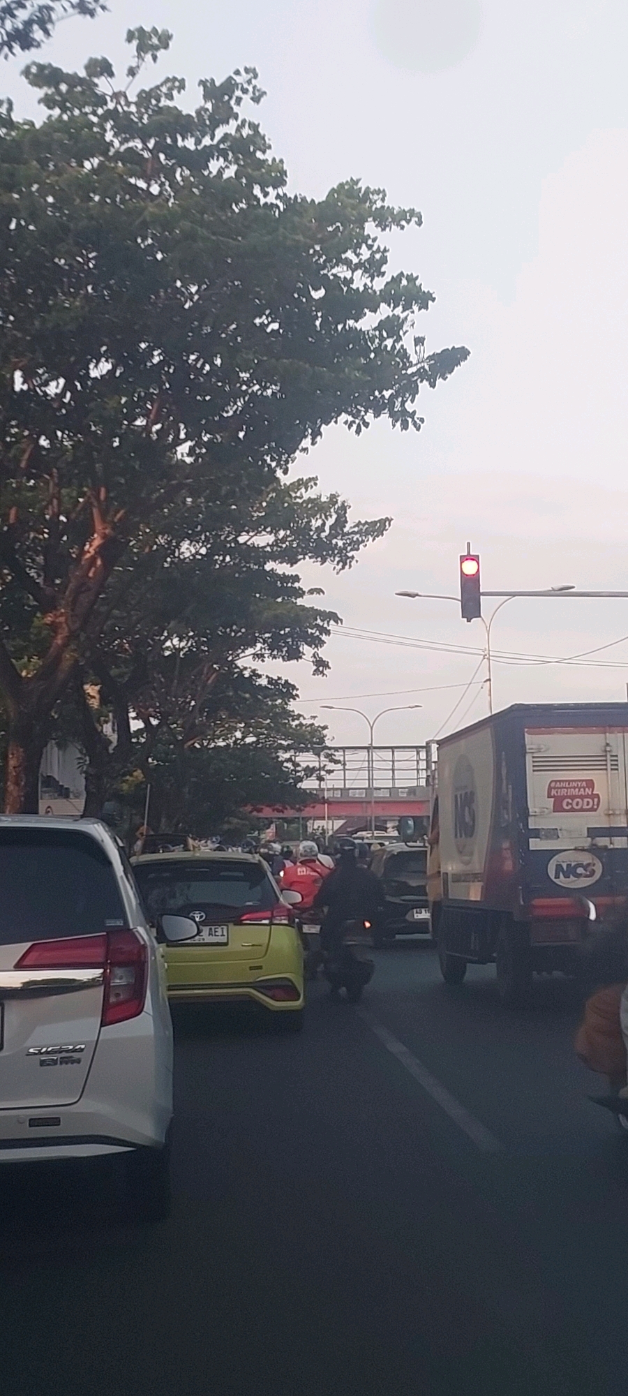 The image depicts a street scene in Surabaya, Indonesia, likely during daylight given the sky's appearance. A traffic jam is evident, with numerous vehicles, including cars and motorcycles, occupying the road. In the foreground, a silver car is partially visible, with the word "SIERRA" on its rear. To its right, a yellow hatchback is seen. A large truck with the NCS logo is also present, featuring text that appears to be Indonesian. A traffic light is illuminated red, contributing to the traffic congestion. Overhead, a pedestrian bridge spans the road. Lush green trees line the left side of the street, with their branches extending into the frame. The sky is overcast, suggesting possibly hazy or cloudy weather. The overall impression is a typical urban traffic scenario in a bustling Indonesian city.