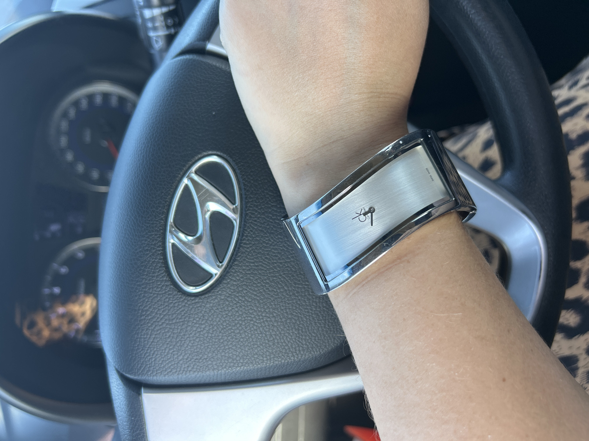 An arm wearing a silver Calvin Klein watch is positioned in front of a car's steering wheel. The steering wheel has a Hyundai logo in the center. The dashboard displays blurry instrument cluster readings. The background is out of focus, showing a hint of patterned clothing. The scene is indoors, likely within a vehicle, with no discernible external environmental cues. The location is not determinable from the provided image.