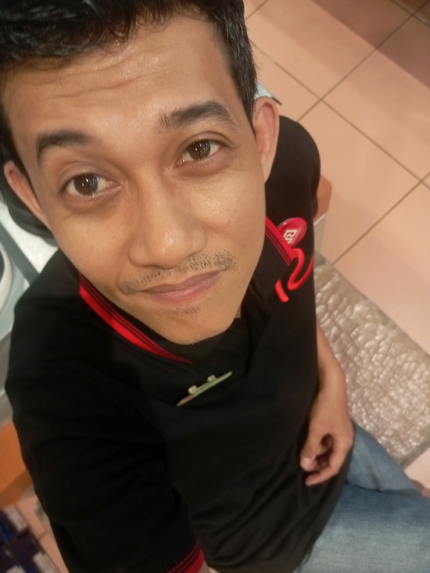 A selfie taken from a high angle shows a man with dark hair and brown eyes. He has a light mustache and stubble on his chin. He is wearing a black t-shirt with a red collar and red graphics on the sleeve, and blue jeans. The man appears to be indoors, with a tiled floor visible in the background. The image was taken in Batam, Indonesia.