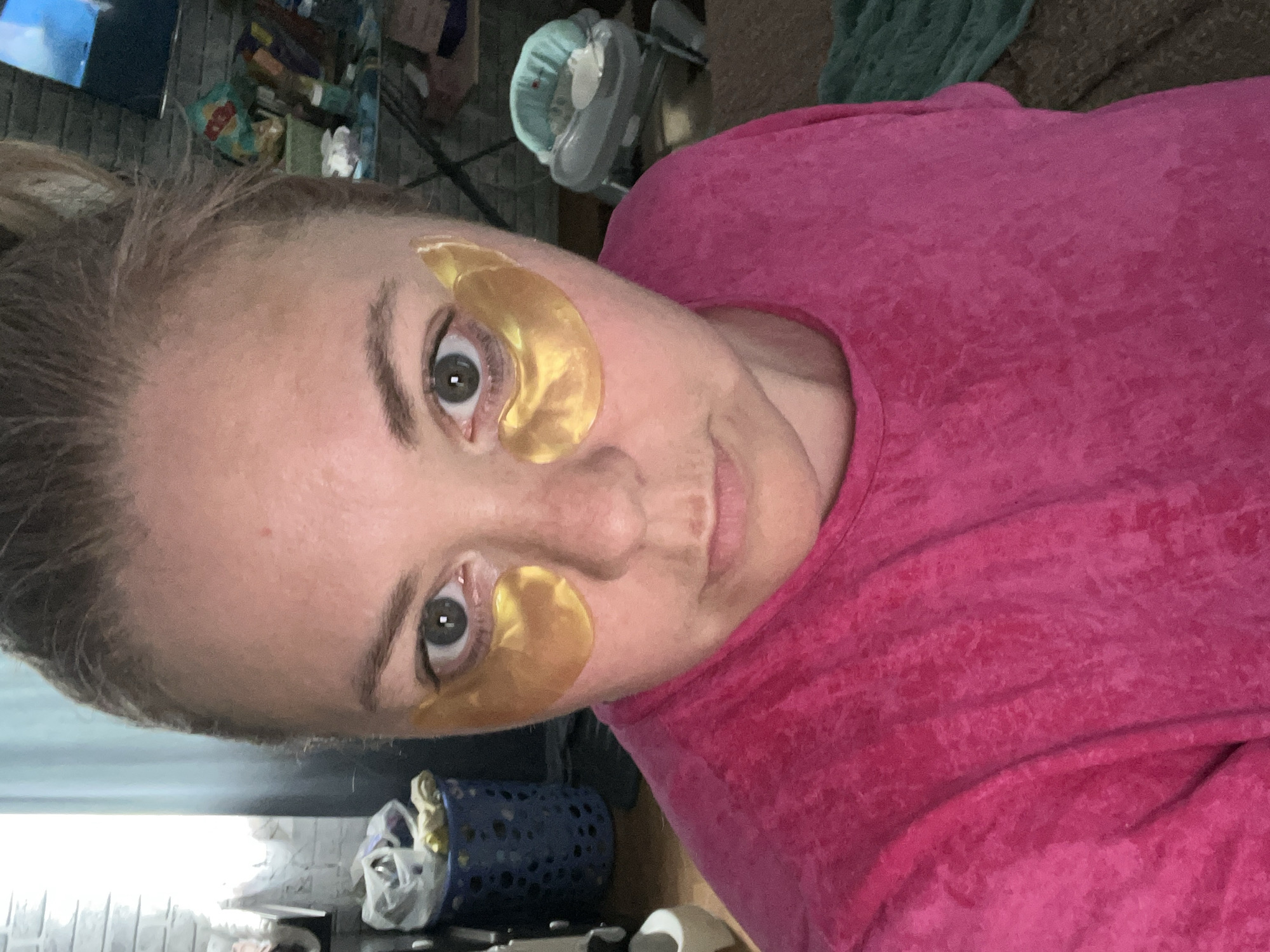 A woman with blond hair pulled back in a bun is wearing pink clothing and gold under-eye masks. The scene contains household items in the background. The city of Vesyoloye, Russia is not depicted.