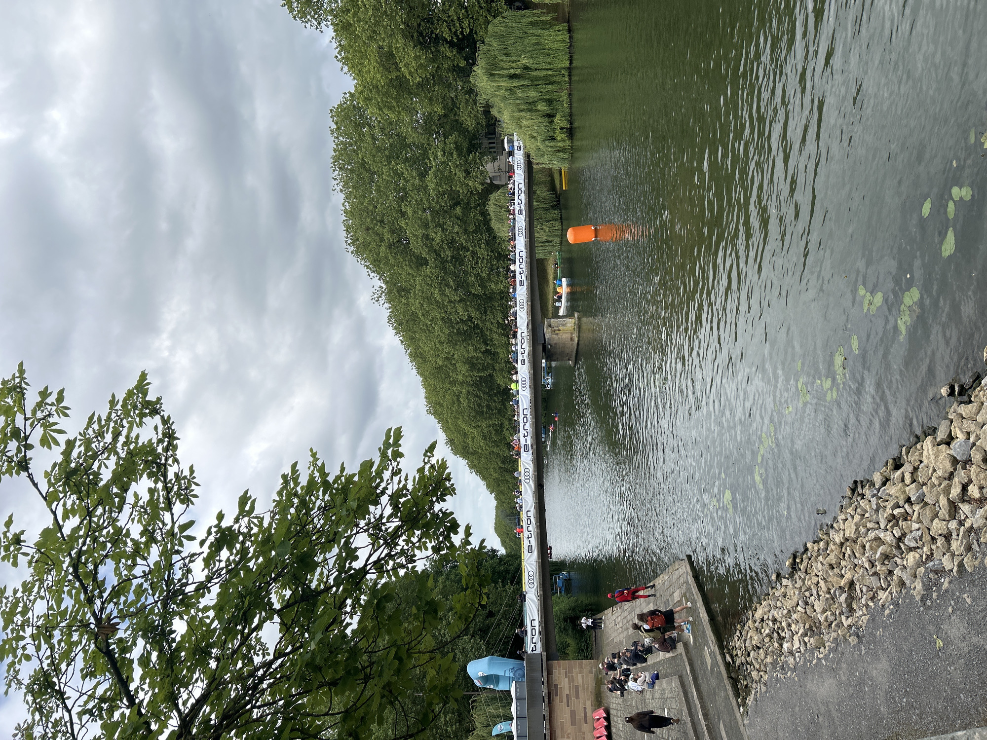 This image captures a view of the Neckar river in Heilbronn, Germany. A bridge spans the river, adorned with a banner featuring the Audi e-tron logo. On the bridge and along its railing, a crowd of spectators is gathered, suggesting an event is taking place. Several boats, including canoes and possibly a larger vessel, are present on the water. An orange buoy floats in the river, marking a section of the course. On the riverbank in the foreground, people are seated and standing on stone steps, observing the scene. The surroundings are lush with trees lining the riverbanks, and the sky is overcast.