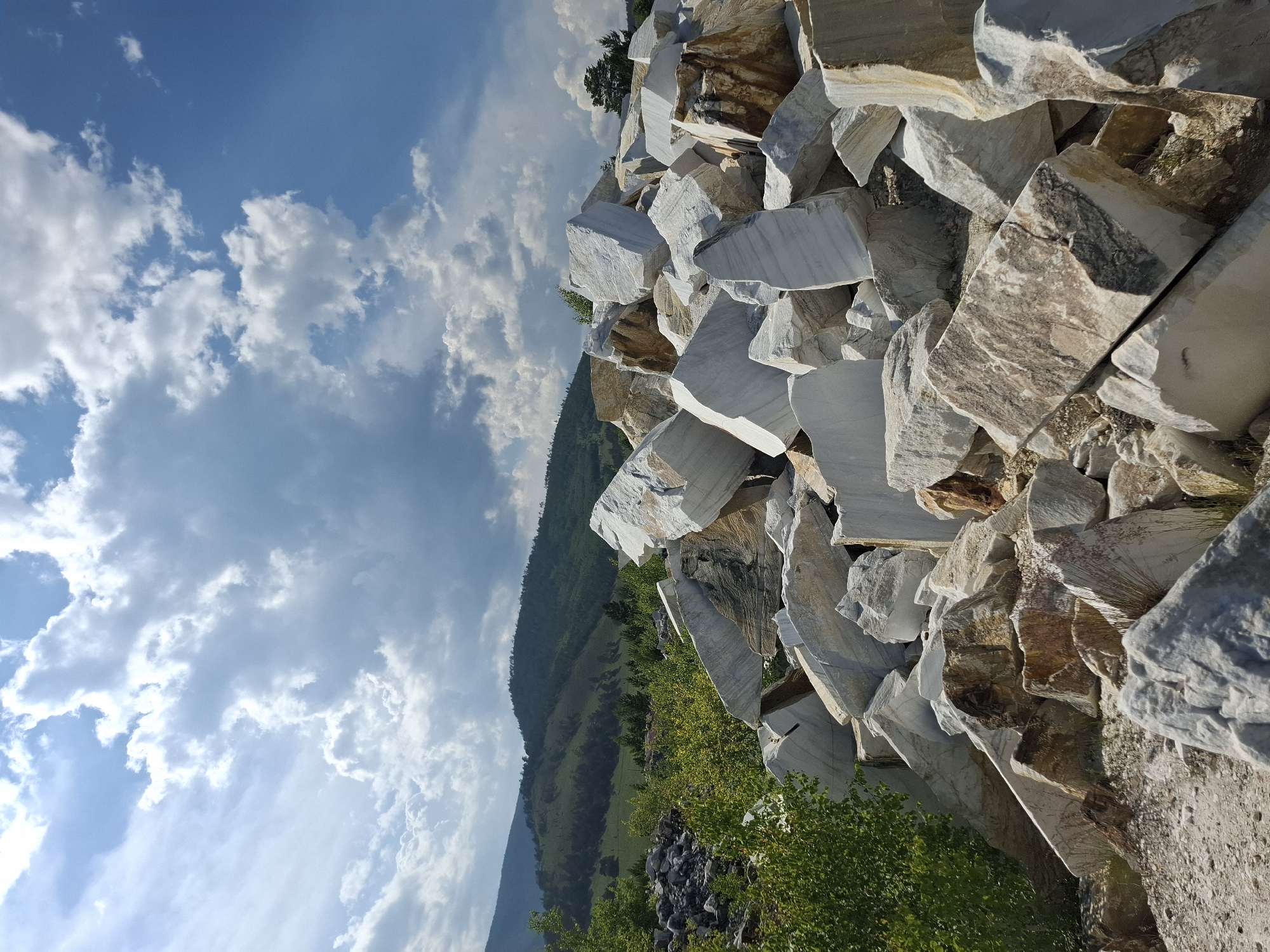 The image depicts a quarry site near Bugul'deika, Russia, filled with large, irregularly shaped blocks of marble. The marble exhibits variations in color, ranging from pure white to shades of gray and brown, with distinct veining patterns. The blocks are piled high in the foreground, suggesting active extraction. In the background, a lush green hillside is visible, dotted with trees and under a partly cloudy sky. The overall scene is one of industrial activity set against a natural landscape.