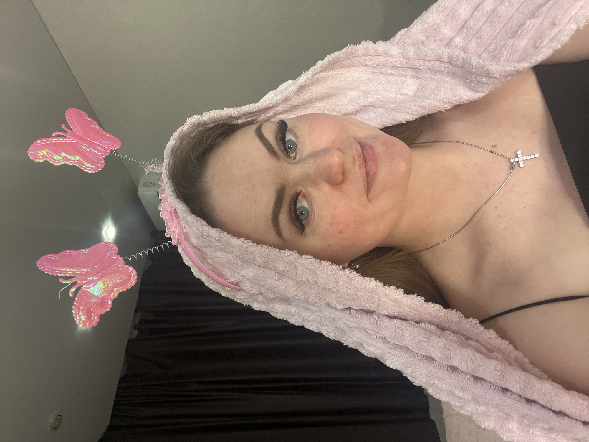 A close-up indoor shot features a woman with fair skin, green eyes, and light brown hair, looking directly at the camera with a soft smile. She has visible false eyelashes and a scattering of freckles on her face. Her head is wrapped in a light pink, fluffy, towel-like material, possibly a spa headband or turban, with subtle pink ribbon details.
Above her head are two prominent, iridescent pink butterfly decorations, each attached to a spring, giving them a whimsical appearance. She wears a silver-colored necklace with a sparkling cross pendant on her neck. A black clothing strap is visible on her right shoulder.
The background is simple and uncluttered, consisting of a plain dark gray wall and a black surface, possibly a bed or curtain. A small, indistinct light source is visible behind the lower butterfly decoration. The lighting appears to be artificial, typical of an indoor setting. No specific location cues are visible within the image itself to suggest Sevastopol, Ukraine. The overall mood is pleasant and playful.