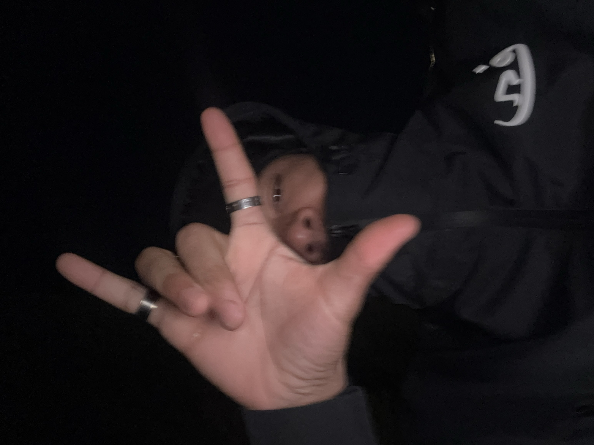 A person is making a hand gesture with their left hand, wearing two rings. The index and pinky fingers are extended, while the middle and ring fingers are folded down. The scene is dimly lit, but it is possible to see a hooded person behind the hand, wearing a black jacket with a white logo on the right shoulder. The setting appears to be outdoors at night, likely in a city, but the exact location within Antony, France is not discernible from the image.