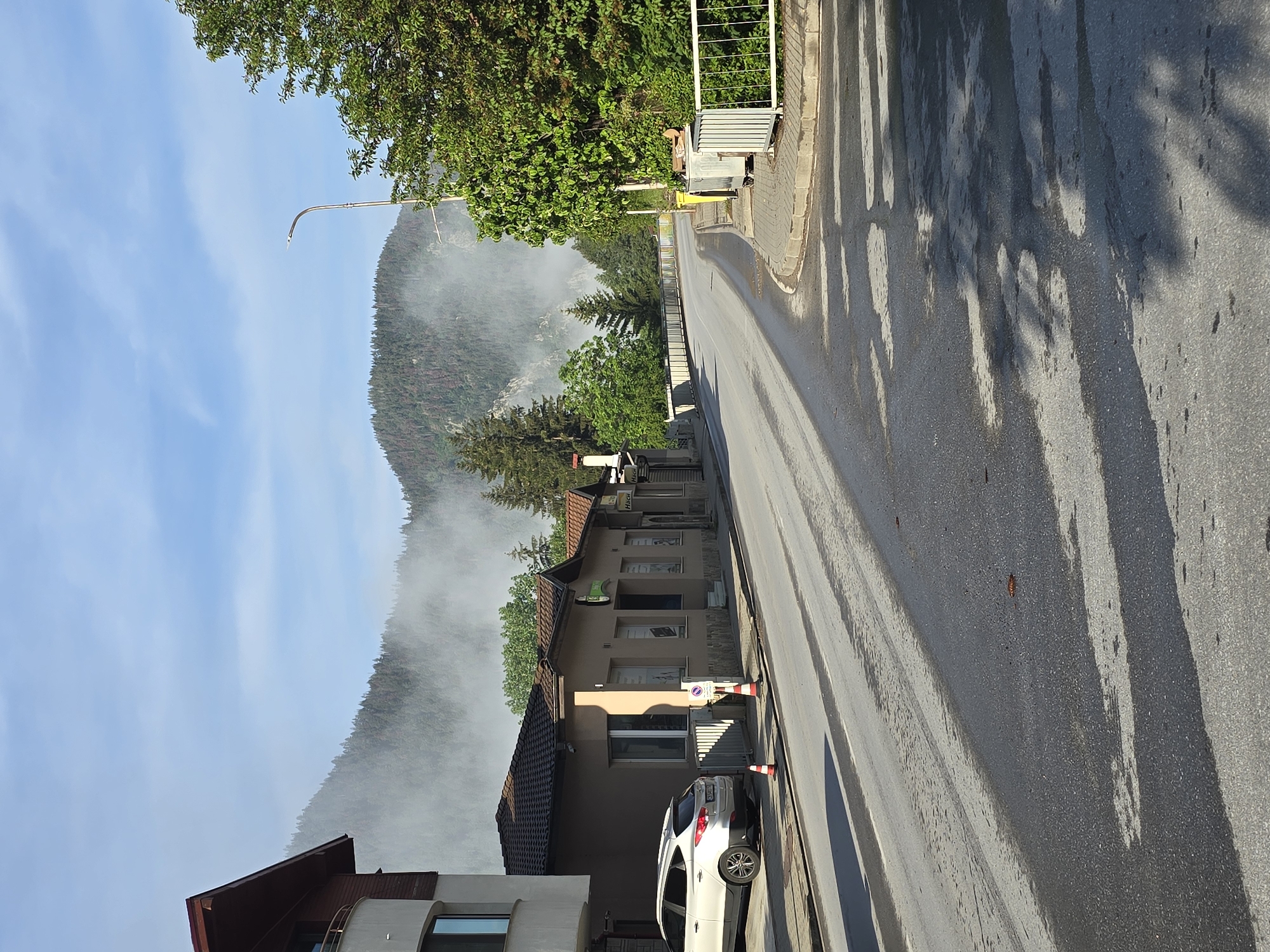 The image displays a street scene set against a backdrop of forested mountains partially shrouded in mist. The perspective looks down a street that curves slightly to the right, with buildings lining its left side and dense trees on the right and in the distance.
**Subjects:** An asphalt road, appearing wet in patches and reflecting light, occupies the right half of the foreground, extending into the distance. Long, distinct shadows are cast across the road by unseen objects to the right, suggesting an angled sun. On the left side of the road, a row of buildings stands. The most prominent is a multi-story structure with warm-toned walls and a brown tiled roof. A white SUV, possibly a Hyundai Tucson, is parked diagonally on the pavement in front of this building, flanked by two orange and white traffic cones. Further down the street, other buildings with multiple windows and commercial facades are visible. Lush green trees line parts of the road and cover the steep slopes of the mountains in the background. A modern, curved streetlight extends into the clear blue sky in the upper left. A light-colored metal fence or gate is visible further down the road on the right.
**Activity or event taking place:** The scene is static with no people visible, suggesting a quiet moment. The parked car and traffic cones indicate a localized arrangement on the pavement.
**Notable details:** The most striking feature is the thick layer of white mist or low-hanging clouds that partially obscures the lower sections of the large, forested mountains in the background, creating a serene and somewhat ethereal atmosphere. Above the mist, the sky is clear and bright blue. The long shadows on the road strongly suggest either early morning or late afternoon. The wet patches on the road could be from recent rain, morning dew, or cleaning.
**Visible text:** On a sign above one of the commercial buildings further down the street, the word "Haci" is partially visible. On a white sign near the traffic cones, a red circular "no parking" or "no stopping" symbol is present, with some small, illegible text underneath.