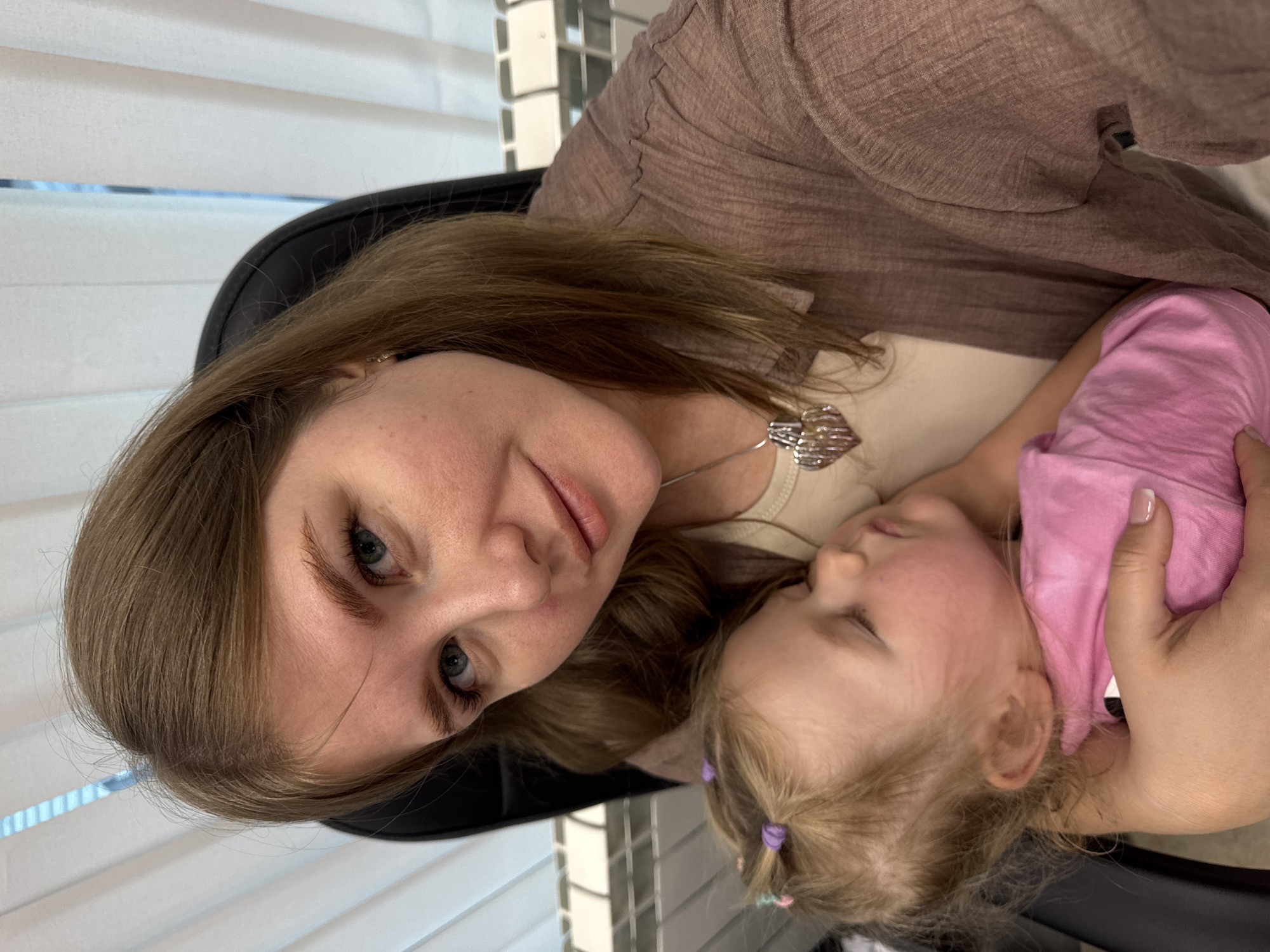 A woman with blue eyes and light brown hair holds a sleeping toddler with blonde hair in her arms. The woman wears a brown, long-sleeved shirt and a silver necklace. The child wears a pink shirt and has two small hair ties in her hair. They are sitting in a black chair with white blinds visible in the background. The location is likely in Perm, Russia, based on the image's origin.