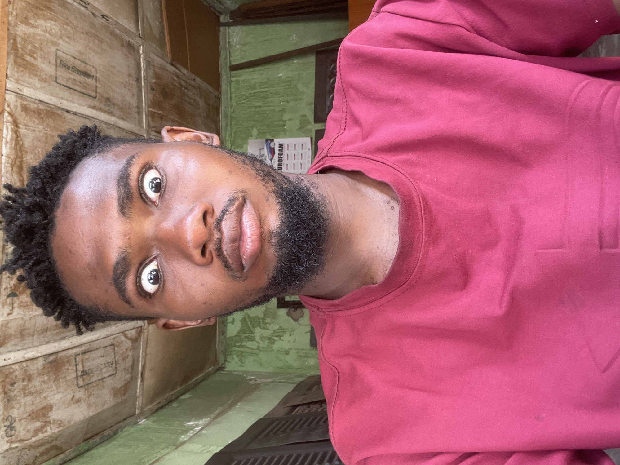 A medium-shot captures a Black male wearing a maroon T-shirt. He has short, curly hair, a dark beard, and dark eyes. The background consists of wooden boxes stacked against a wall and a green wall. A calendar is also visible. The location is Zaria, Nigeria.