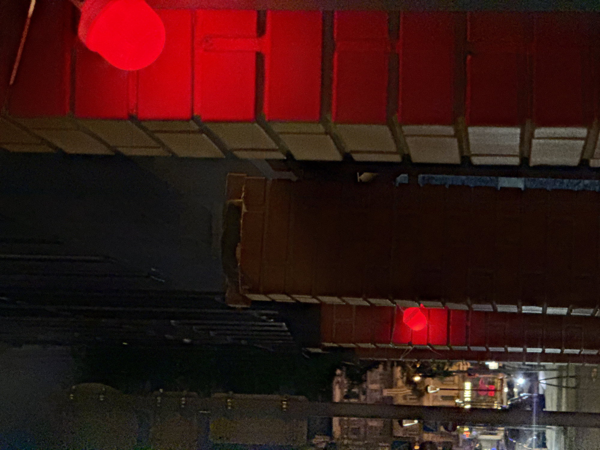 The image shows a night scene in Moscow, Russia, captured from a high vantage point. The focus is on the underside of a red metal structure, possibly a bridge or a portion of a building's facade. The structure is illuminated with red lights. Below this, a dimly lit street is visible with various buildings and streetlights. The overall impression is a view of the urban landscape at night, dominated by the prominent red structure.