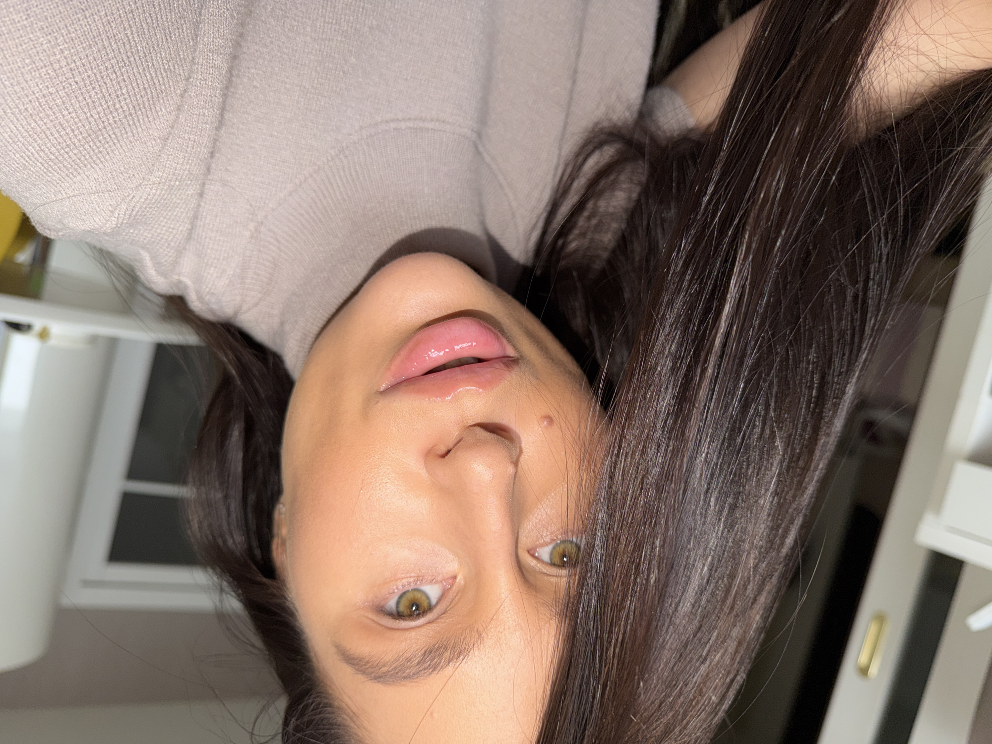 This is a close-up, rotated portrait of a person, likely female, situated in an indoor setting. The image is oriented sideways, with the person's head appearing upside down, tilted towards the bottom of the frame, and their body extending upwards.
The main subject is a person with long, dark brown hair that cascades across the frame. They are wearing a light beige or taupe ribbed knit garment, possibly a sweater or top. Their face is well-lit, showing prominent light-colored eyes, appearing hazel or light green, which look directly at the viewer. Their lips are full and have a glossy, light pink finish. The skin appears smooth, suggesting makeup application. A small mole or blemish is visible on the bridge of the nose.
The activity taking place is likely a selfie or a close-up portrait session. The person's expression is neutral to subtly serene.
The background is softly blurred, indicating an indoor setting. Visible elements include light-colored furniture or architectural features, such as what appears to be part of a white window frame or a white shutter on the right side of the image, and a white, possibly shelf-like structure on the left. The lighting is soft and consistent with an indoor environment, making it difficult to ascertain the exact time of day. No specific weather conditions are discernible. There is no visible text in the image. The location context provided is Khimki, Russia.