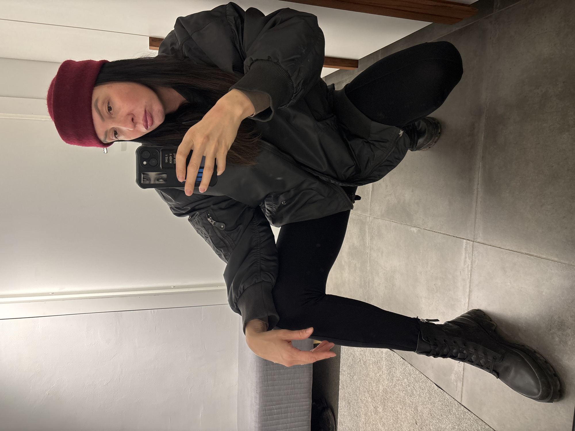 A woman with long dark hair and a maroon beret is squatting in a room with gray tile floors and light gray walls. She is wearing black leggings, black boots, and a black bomber jacket with a zippered pocket on the left sleeve. She is holding a smartphone in her left hand, taking a selfie. The reflection in the phone shows a partial view of her face and the surrounding environment. A wooden beam is visible near the top of the frame. The scene appears to be indoors. The location in the city of Chaikovsky, Russia, cannot be confirmed from the image.