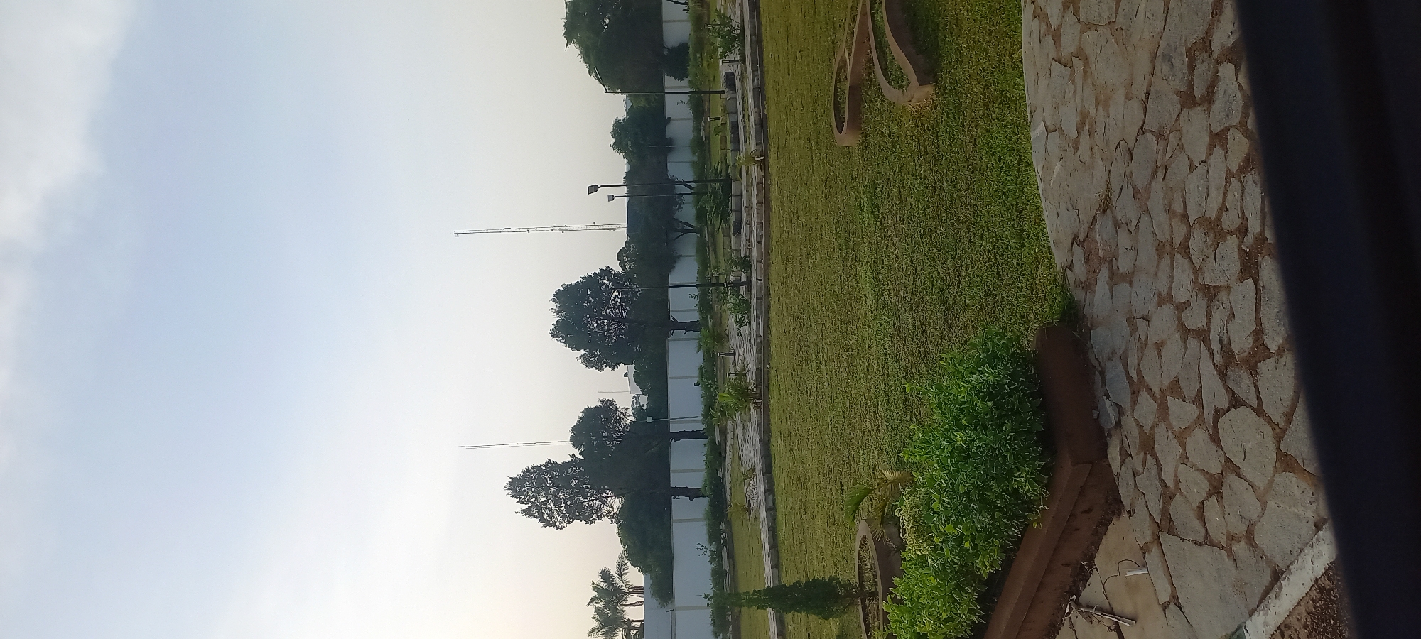 The image depicts an outdoor scene in Barikin Jirga Summa, Nigeria. In the foreground, a pathway paved with irregular flagstones leads into a well-maintained lawn area. To the left, a raised planter bed contains lush green shrubs. Beyond the lawn, a paved walkway winds through the grassy area, bordered by low hedges and ornamental plants. Several streetlights are visible along this walkway. In the background, a white wall with a fence runs horizontally across the image, with various trees and palm trees planted behind it. Two tall communication towers stand prominently against the pale sky, one taller than the other. The overall scene suggests a public park or a well-landscaped area within a city. There are no people visible in the image.