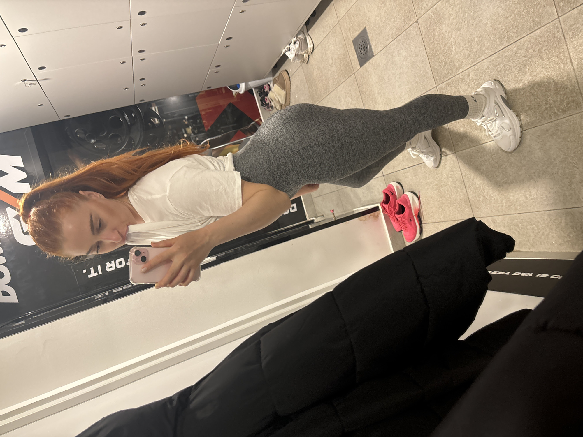 The scene is set in a gym locker room located in the city of Budva, Montenegro.
A female individual with long, reddish-blonde hair tied in a high ponytail is depicted taking a mirror selfie. She is wearing a white short-sleeved t-shirt, form-fitting grey leggings, and white athletic shoes with white socks. Her right hand holds a light pink smartphone, encased in clear plastic, pointed towards her reflection. Her body is angled slightly, presenting a partial profile view, with her head turned towards the phone. Her lips are slightly puckered.
The background consists of a dark wall with large white text, partially legible as "FOR IT." and "GYM." To the right of the subject, a series of white lockers with multiple compartments and dark circular fasteners are visible. On the left, a black padded jacket hangs. The floor is covered with light-colored square tiles, featuring visible grout lines and a square metal drain cover. Situated on the floor near the lockers are a pair of bright pink athletic shoes, a pair of light-colored slippers, and a partially visible white waste bin. The environment is uniformly illuminated by artificial indoor lighting.