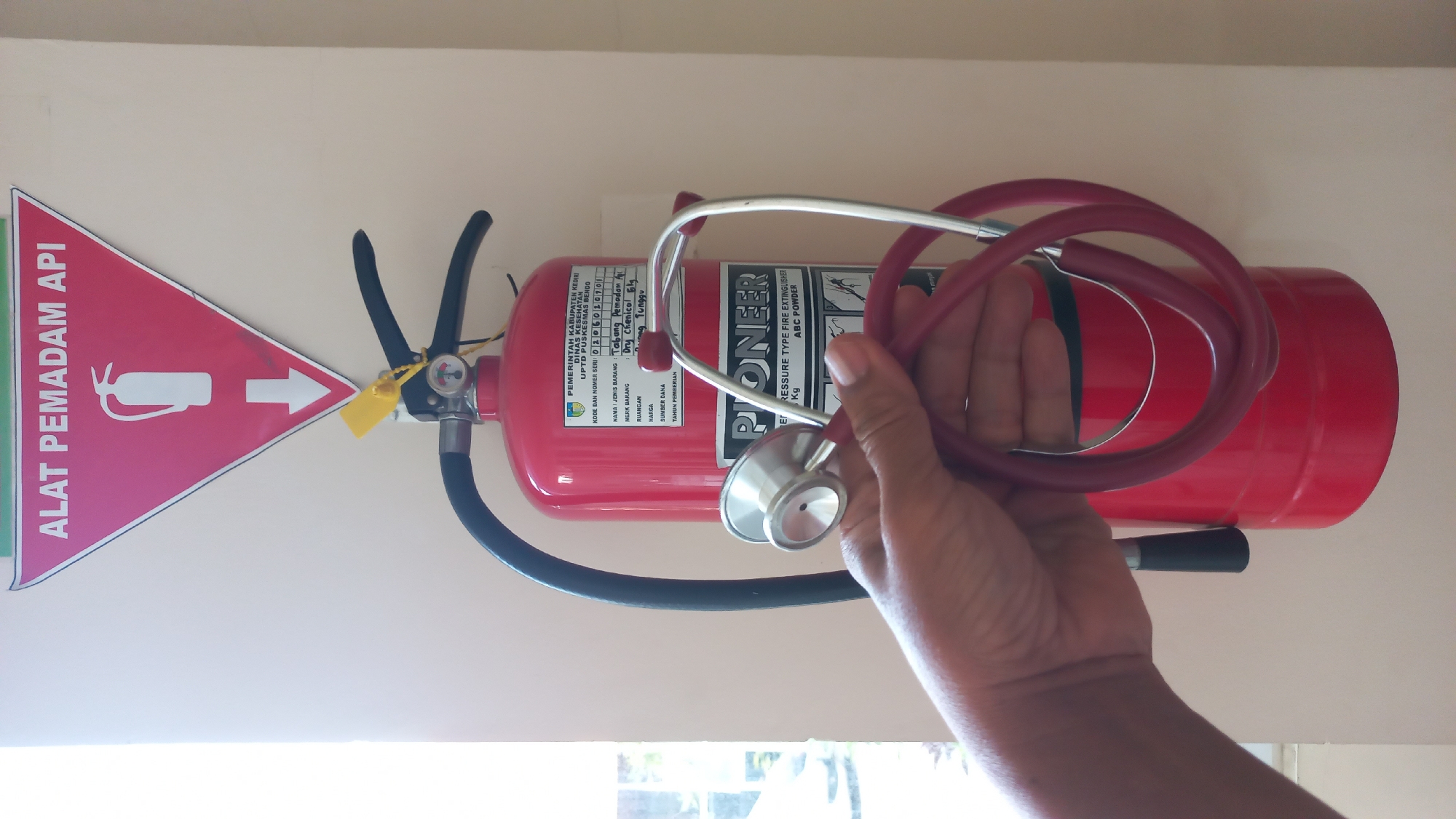 In the city of Pare, Indonesia, a red fire extinguisher is mounted on a light-colored wall. Above the extinguisher, a red triangular sign with a white outline displays the text "ALAT PEMADAM API" (Fire Extinguisher) in white lettering, and a white icon of a fire extinguisher with a downward-pointing arrow. A yellow tag is attached to the top mechanism of the fire extinguisher. A person's hand is holding a red stethoscope in front of the fire extinguisher. The stethoscope's chestpiece and tubing are visible. The fire extinguisher has a label that reads "PIONEER" and indicates it is a "PRESSURE TYPE FIRE EXTINGUISHER" with "ABC POWDER."