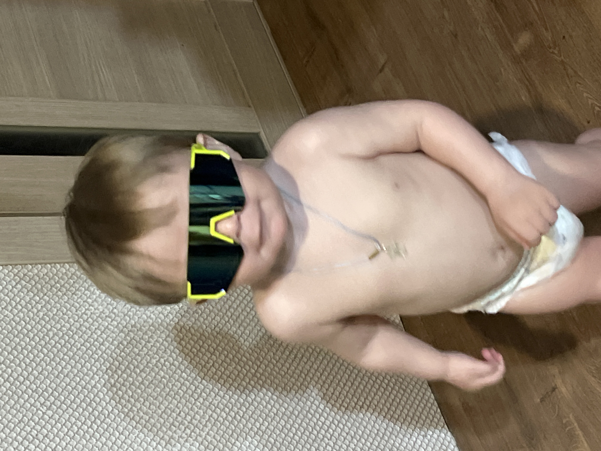 A toddler wearing a diaper and large sunglasses lies on the floor, which has both carpet and hardwood sections. The carpet section is beige and textured. The hardwood section is brown. The toddler's torso is visible, and they are wearing a necklace. A light-colored door or wall trim is visible in the background to the left. The location in the city of Noviy, Russia cannot be determined from the provided image.