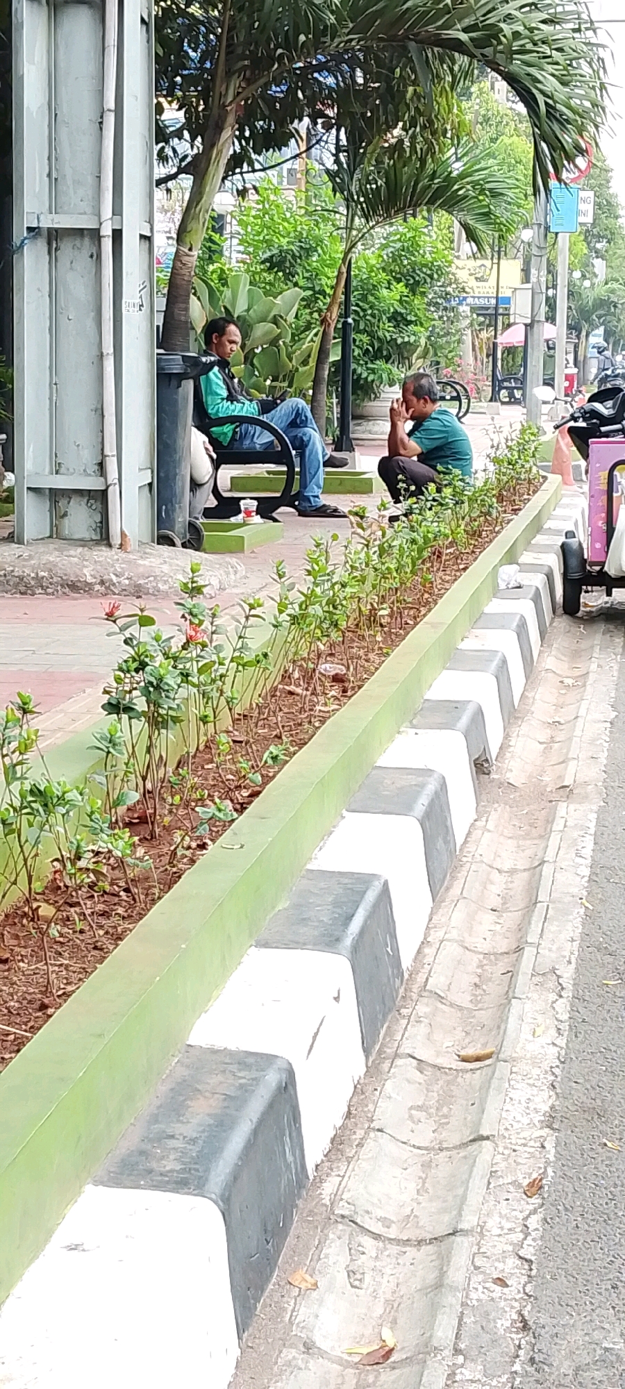 The image captures an outdoor scene in the city of Bekasi, Indonesia, likely a public space with a sidewalk and a road. In the foreground, a long planter box with young green plants separates a paved pedestrian area from a concrete drainage ditch. The planter box has a light green border. Beyond the planter, two men are visible. On the left, a man sits on a dark bench, facing away from the camera, wearing a teal jacket and blue jeans. He appears to be looking at a phone. To his right, another man is crouched down, facing the camera, wearing a dark green t-shirt and dark pants. He has his hands clasped in front of his face. In the background, there are trees, foliage, and some urban infrastructure including streetlights and signs. A small pink and white cart with wheels is parked to the right of the image, and a motorcycle is partially visible behind it. The overall impression is of a casual, everyday moment in a public urban setting.