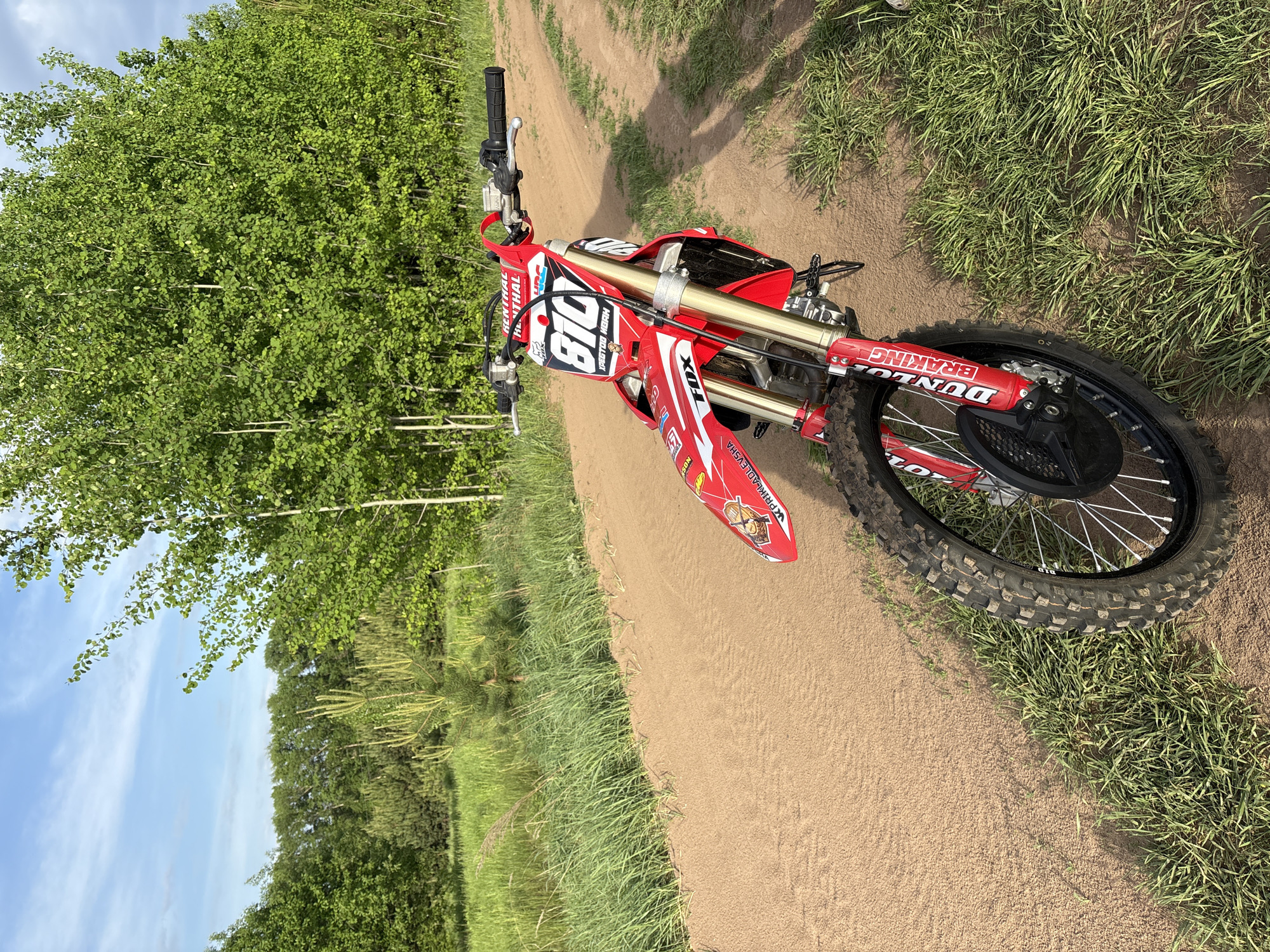 A red and white dirt bike is parked on a dirt path next to a grassy area with a dense green tree line. The sky is blue with some clouds. There is no indication of the location being in the city of Votkinsk, Russia or any presence of people.