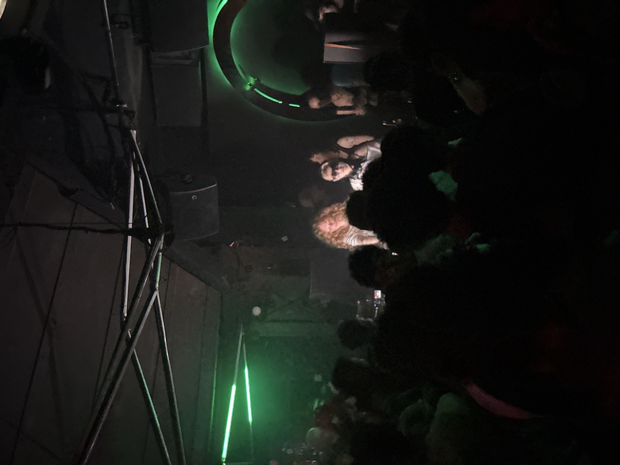 The image captures an indoor nightclub or event space in Paris, France, illuminated predominantly by green light. A crowd of individuals occupies the foreground, their heads and upper bodies appearing as dark silhouettes, facing an elevated platform or stage. On this stage, three primary figures are discernible. To the left, a person with long, wavy light-colored hair stands behind what appears to be DJ equipment. To their right, a second individual with short, light hair wears sunglasses and a light-colored top, with headphones positioned around their neck; this person faces left. Further to the right on the stage, a third individual with an arm tattoo is partially visible in dim light, facing generally right. Overhead structures include metal trusses, cables, and mounted dark speaker enclosures. The background on the right features a large, vertically oriented circular archway emitting green light from its inner rim. Horizontal green strip lights are also visible on the upper left side of the scene. The overall environment indicates an active live music or DJ performance.