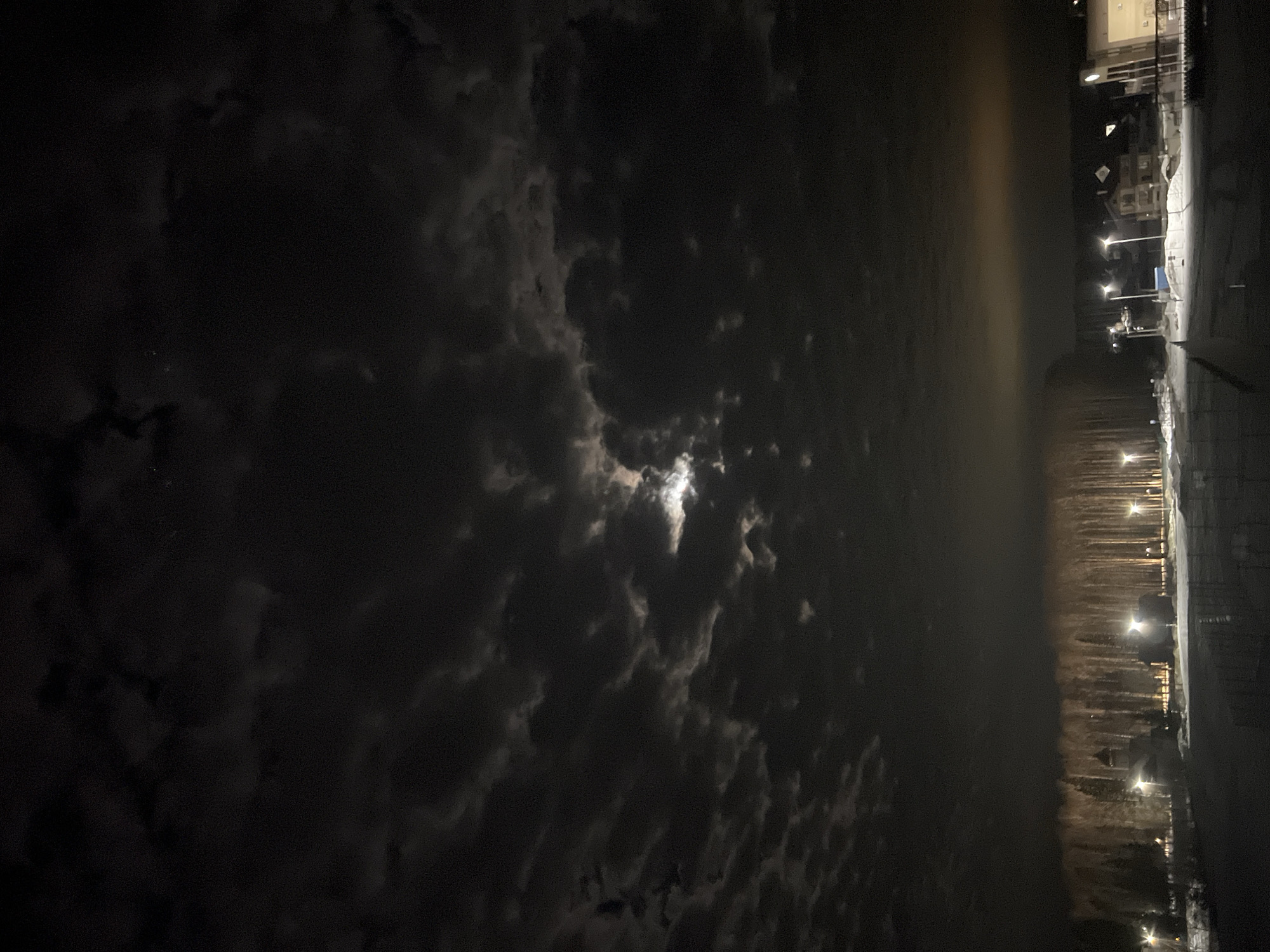 A night scene shows a partially obscured moon through a cloudy sky. The right side of the image includes an urban setting with illuminated streetlights, snow-covered ground, and buildings. The location is near Noviy, Russia.