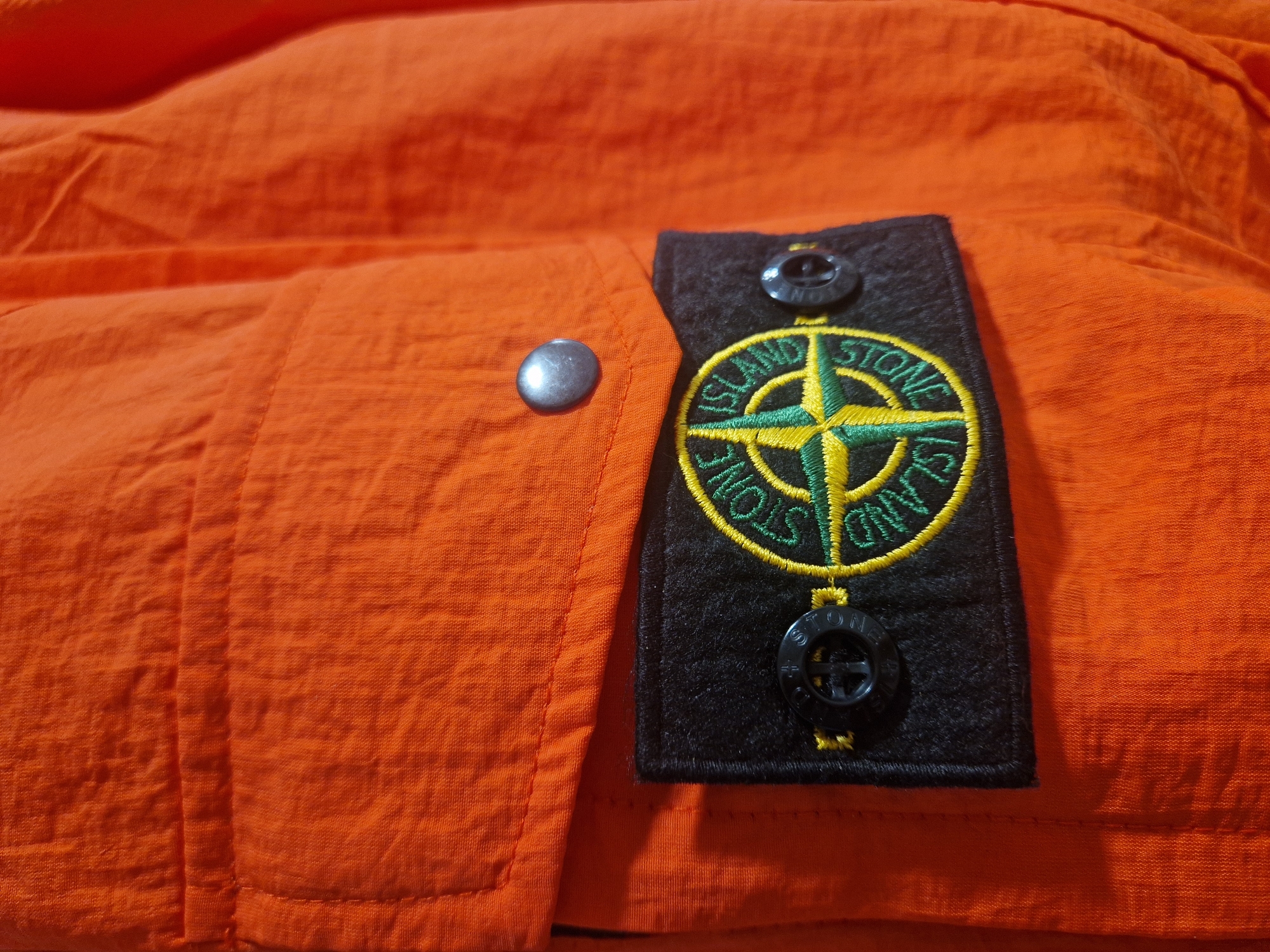 A close-up shows an orange garment with a Stone Island logo patch attached. The patch is black with a circular emblem featuring a green and yellow compass rose, surrounded by the words "STONE ISLAND". Two black buttons secure the patch. A single silver button is visible on the orange fabric adjacent to the patch. The texture of the orange fabric appears wrinkled and slightly translucent. The location is Unknown, Unknown.