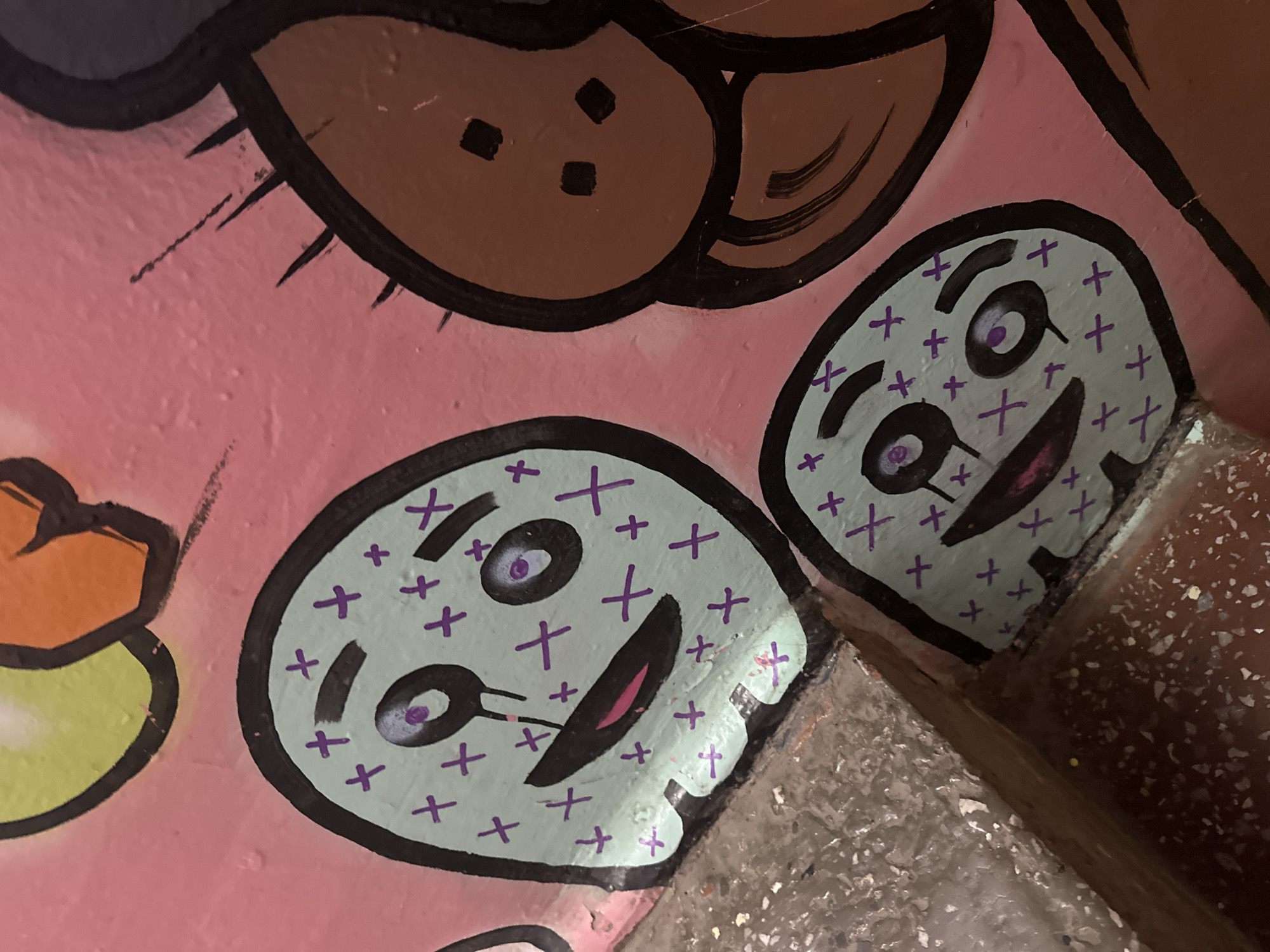 The image shows a section of a wall with graffiti art. The graffiti features two cartoonish, light blue ghost-like figures with purple crosses for decoration, large black eyes with purple irises, and black mouths with pink interiors. One ghost is larger and more prominent, with a tear-like black line extending from its eye. To the right of the ghosts is a brown, bulbous shape with black dots. The background is a solid pink color. In the foreground, there is a textured, gray surface with a rough, chipped appearance, possibly a ledge or the corner of a structure. The overall scene appears to be an outdoor environment, likely a public space with graffiti. The city of Biysk, Russia is mentioned, but there are no specific visual cues within the image to confirm this location.