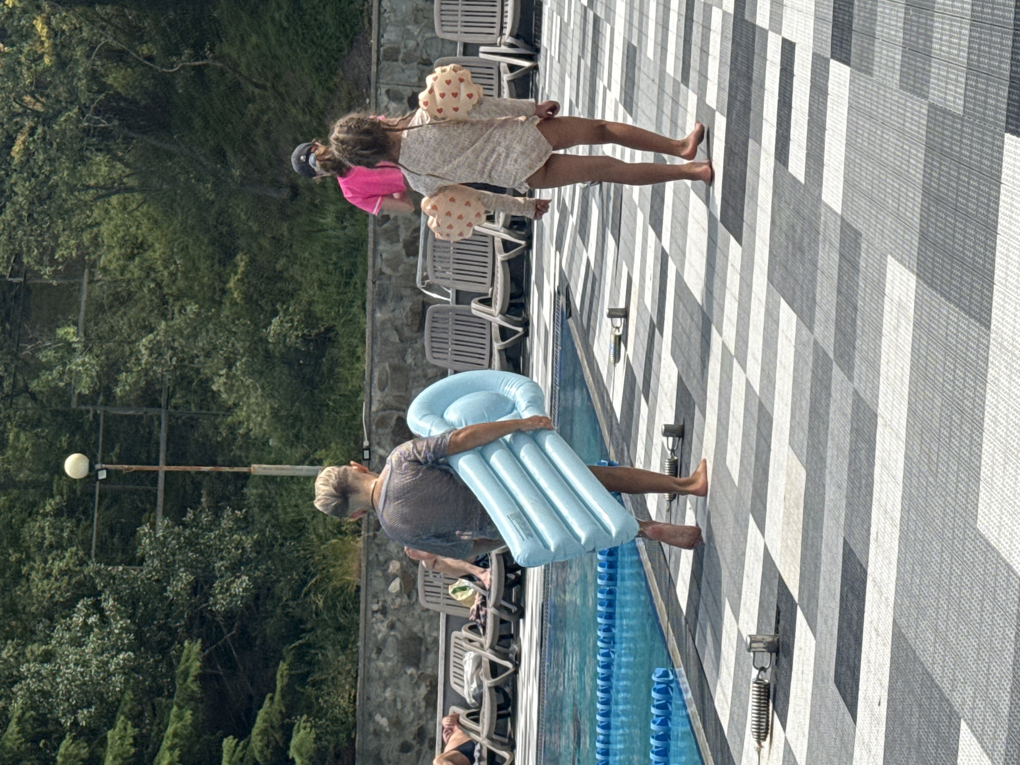 The image depicts a poolside scene in Массандровский поселковый совет, Ukraine. In the foreground, a person with short, styled blonde hair is walking away from the camera, carrying a large, deflated light blue inflatable mattress. The person is wearing a sheer, short-sleeved shirt and appears to be barefoot. To the right, a young girl with braided hair is also walking away from the camera. She is wearing a one-piece swimsuit with a floral pattern and arm floaties adorned with heart shapes. Behind them, another person in a bright pink t-shirt and a baseball cap is visible. The ground is paved with a checkered pattern of grey and white tiles. The background shows a swimming pool with blue lane markers, several lounge chairs, and lush green trees, indicating an outdoor setting.