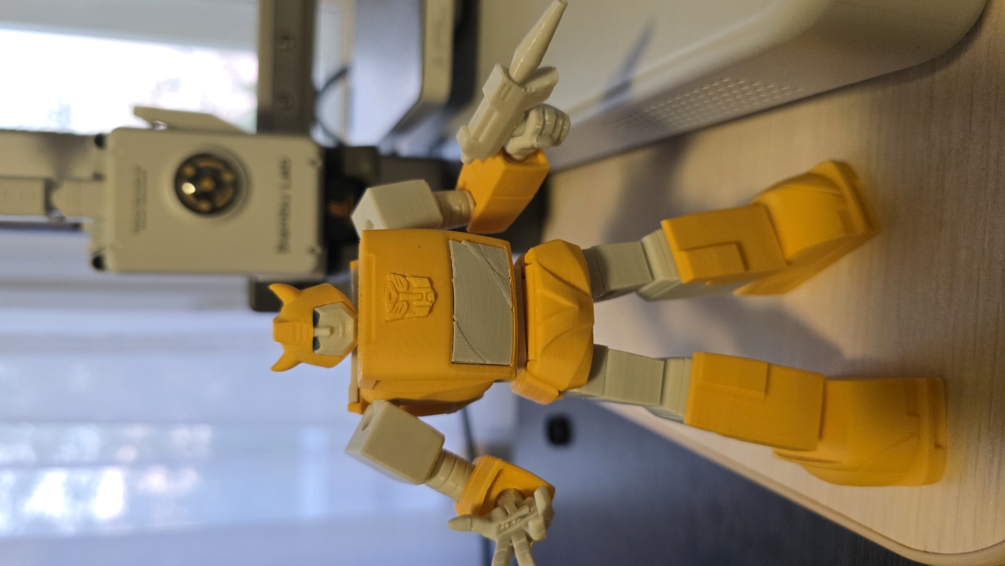 A yellow and white Transformer action figure stands on a wooden surface. The figure is in a humanoid pose with its arms slightly out. Its left hand is held up, with the index and middle fingers extended in a peace sign. Its right hand holds a white blaster accessory. The figure has a yellow helmet with horns, a white face with blue eyes, a yellow chest with a gray Autobot insignia, and yellow legs. In the background, a white and gray machine with a circular lens is visible, as is a blurred image of what appears to be a window with curtains. The scene is lit with soft, artificial light. The image is taken from a slightly low angle, looking up at the action figure.
