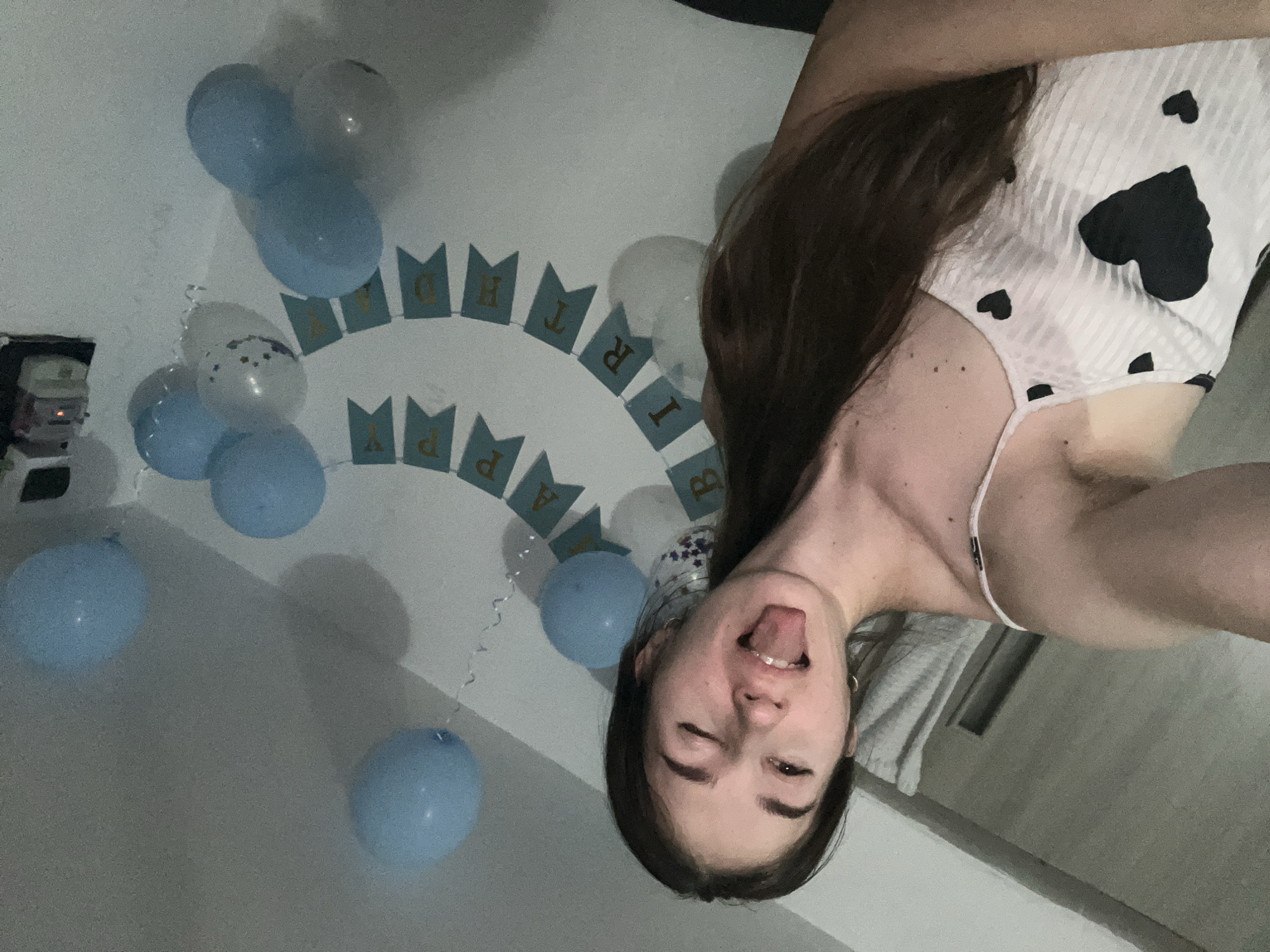 A young woman with long brown hair and light skin is pictured in a selfie. She is wearing a white camisole with black heart patterns. Her left eye is winking and her tongue is sticking out. She is positioned in the foreground, with a wall decorated with blue balloons and a "Happy Birthday" banner in the background. The lighting appears to be dim and artificial. The location is an indoor setting, possibly a bedroom, in an unknown city and country.