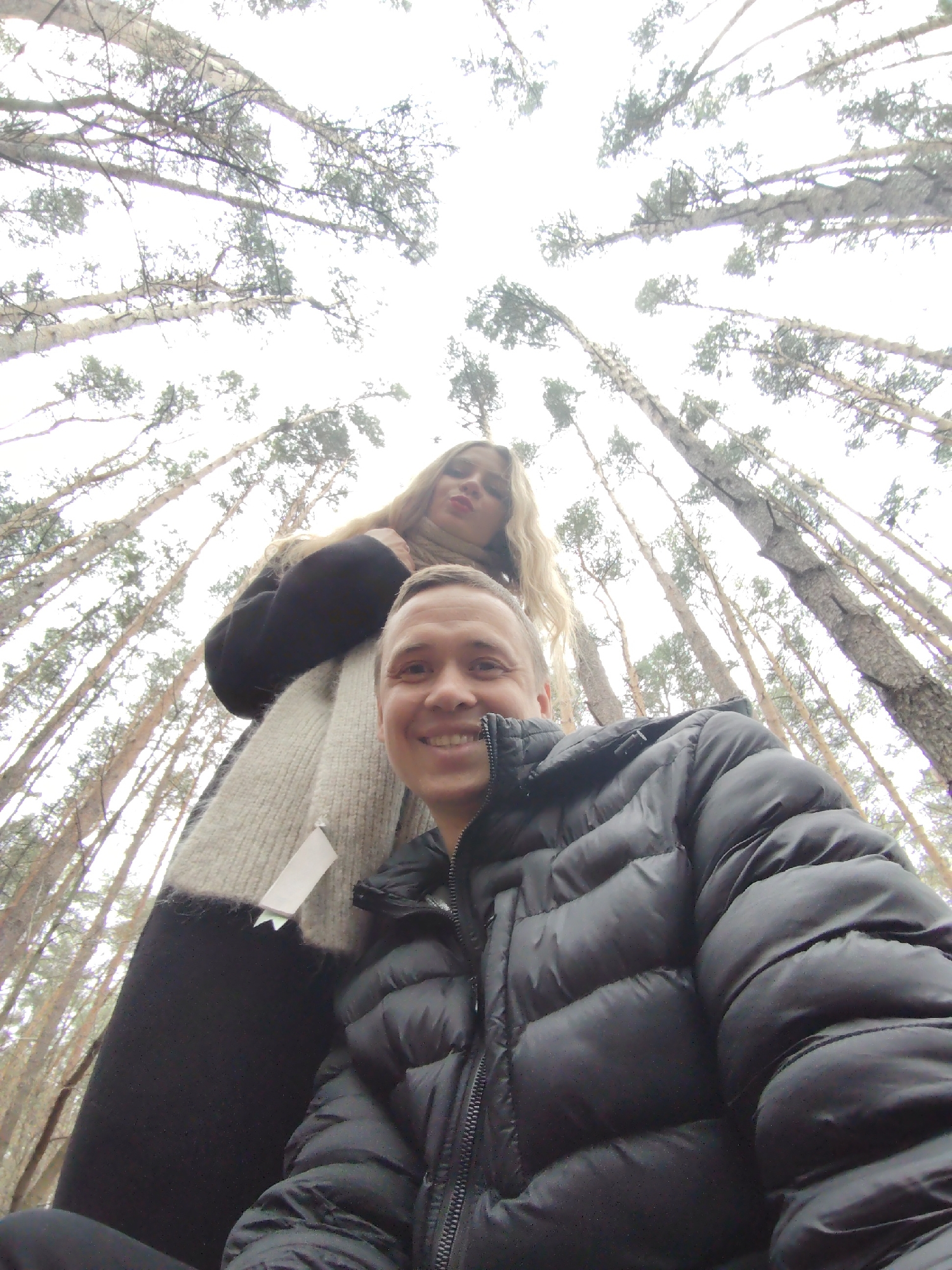 A low-angle selfie shows a man and a woman standing in a forest, the trees towering above them. The man is in the foreground, smiling at the camera, and wearing a black, puffy jacket with a visible zipper. The woman is behind him, looking slightly down and to the left. She has long blonde hair and is wearing a black coat over a light-colored scarf. The camera angle emphasizes the height of the trees that surround them. The trees are thin, with sparse branches, and the sky is visible between them. Based on the location provided, the image was taken in the city of Chaikovsky, Russia.
