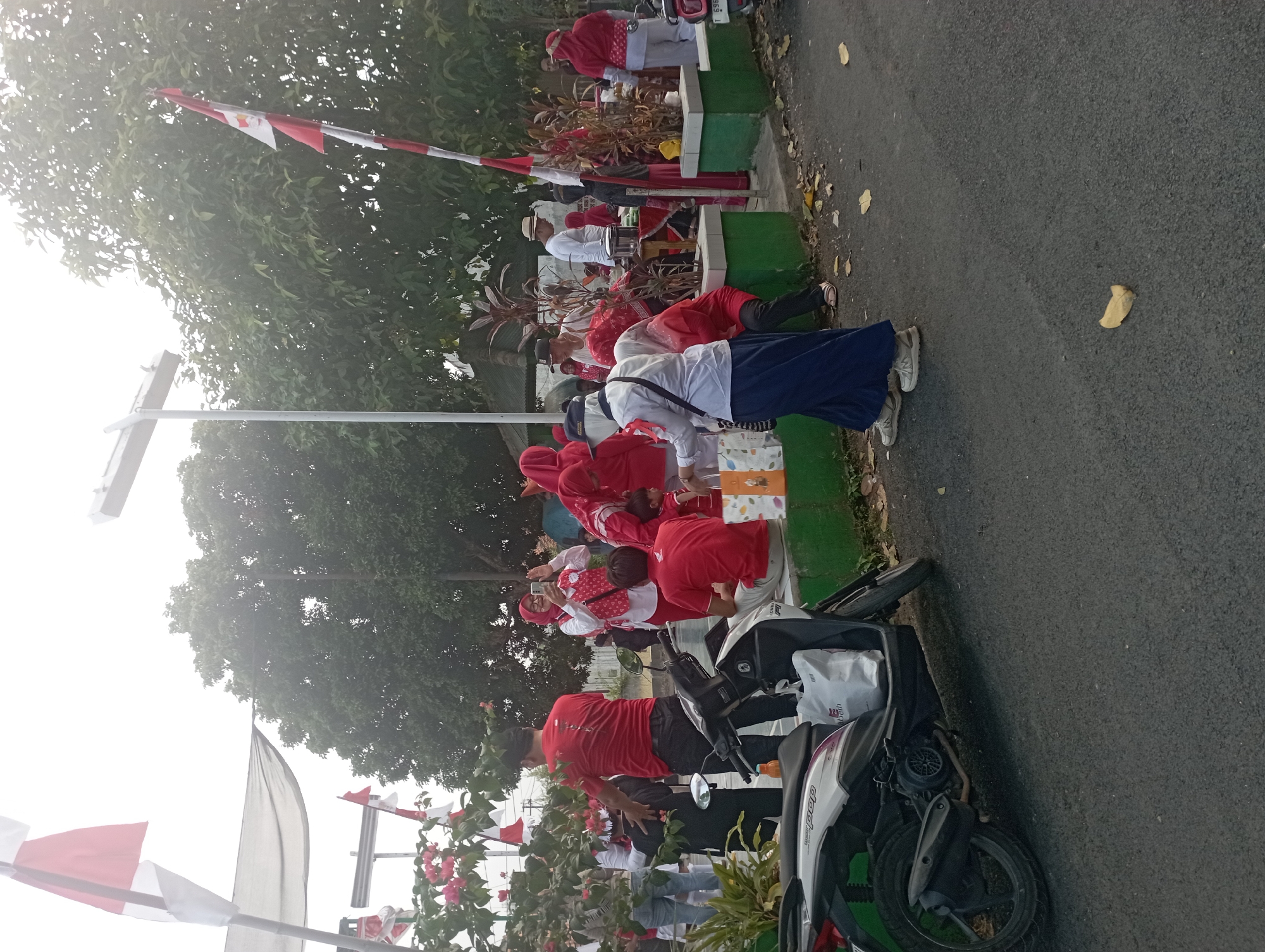 The image depicts an outdoor scene, possibly a public gathering or event, in Purwakarta, Indonesia, with a strong presence of red and white decorations, suggesting a national celebration. In the foreground, a white scooter is parked on the side of a paved road, with a white shopping bag on its seat. Several individuals are present, many wearing red shirts and headscarves. Some people are standing, while others are seated on a low green wall. One woman in the center, wearing a white shirt and blue skirt, is holding shopping bags. Another woman, wearing a red headscarf and a red and white patterned vest, is taking a photo with her phone. In the background, trees are visible, and Indonesian flags are prominently displayed on poles. The overall atmosphere appears to be lively and celebratory.