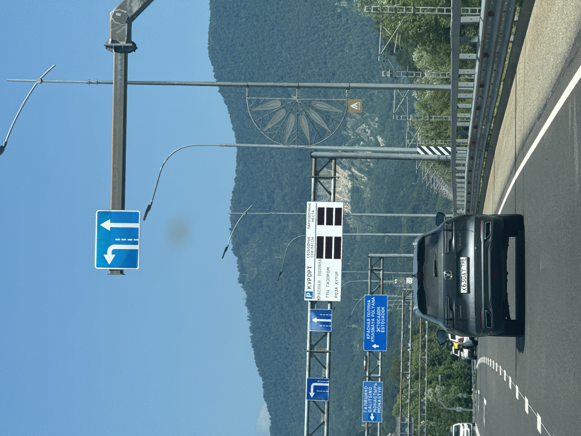The image shows a view from a vehicle traveling on a highway in Adlerskiy, Russia, with lush green mountains in the background. Multiple road signs are visible, directing traffic. One sign indicates that the left lane is for turning left, and the right lane is for going straight. Another sign points to "Galitsino," "Gralitsino," and "Monastery." A third sign directs drivers to "Krasnaya Polyana" and "Estosadok." Further down the road, a larger sign indicates "Resort" with information about parking and specific destinations like "GTC Gazprom" and "Roza Khutor." The road is paved and has white lane markings. A dark-colored SUV with a license plate reading "X630AT 198" is in the foreground, and other vehicles are visible ahead on the highway. The sky is clear and blue. There are also streetlights and electrical infrastructure along the roadside.
