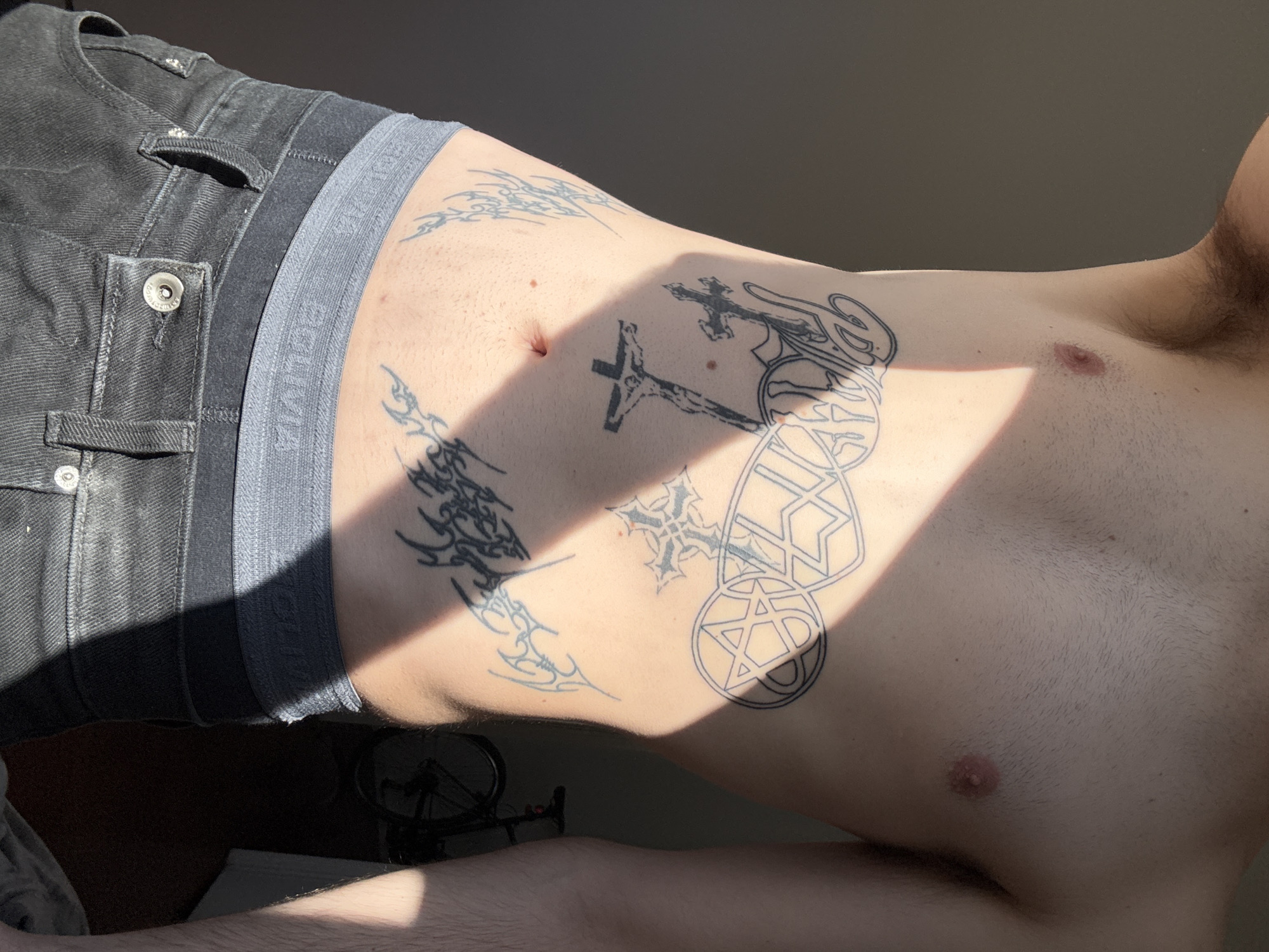 A cropped, vertical shot depicts the midsection and upper torso of a person, likely male, lying down or leaning back indoors. The individual wears black denim jeans or shorts and grey boxer briefs with an elastic waistband. The waistband bears the text "BOLIVIA" in white letters.
Multiple tattoos are visible on the torso. On the left side of the abdomen, there are several tribal-style designs. Closer to the center, a prominent crucifix tattoo features the figure of Christ. To the right of the crucifix, another stylized cross design is present. Further right on the abdomen and ribs, various geometric outline tattoos include a pentagram within a circle, and other abstract shapes. Body hair is visible on the chest and abdomen, along with a few moles.
The scene is illuminated by strong, directional sunlight, creating a distinct diagonal shadow across the body, partially obscuring some tattoos and casting a dark area in the lower left corner. In this shadowed area, parts of a bicycle, specifically a wheel and handlebars, are visible. The background appears to be a plain, dark surface or wall. The bright sun suggests it is daytime, possibly morning or late afternoon. No specific location cues are visible in the image itself, though the provided context is Buenos Aires, Argentina.