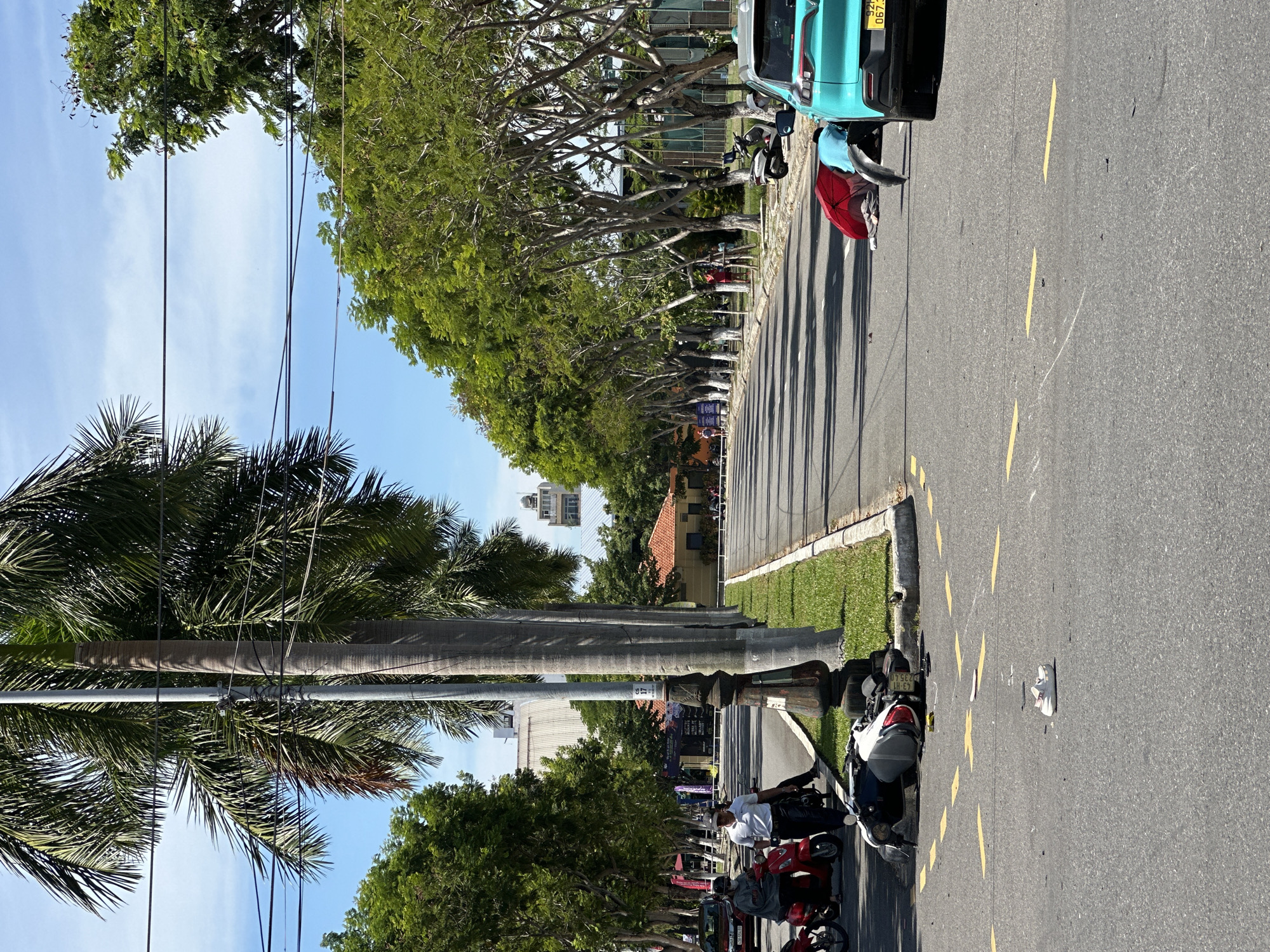 This is a street scene in Da Nang, Vietnam, likely after a traffic accident. In the foreground, a motorcycle is lying on its side on the asphalt road, with scattered debris around it. A single white sneaker is also visible on the road. Further down the road, a turquoise car is parked, and near it, a person is lying on the ground partially covered by a red umbrella, with another person in a blue shirt tending to them. To the left, two people are standing next to a red scooter, and a man in a white polo shirt and dark pants is walking. Palm trees line the right side of the road, and lush green trees are visible on both sides. In the background, buildings and power lines are present. The sky is clear and blue.