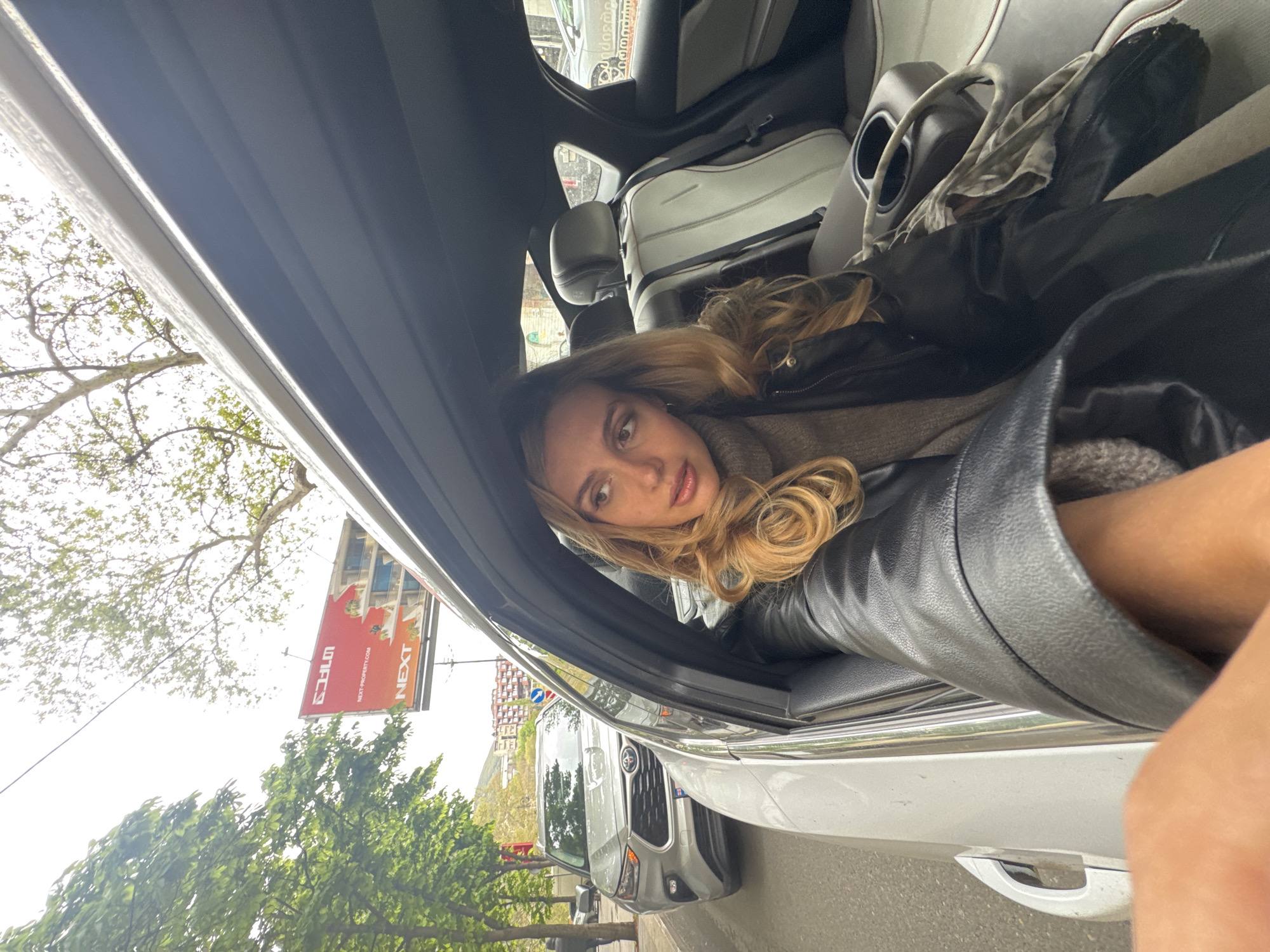 The image is oriented sideways, depicting a young woman leaning out of a car window. She has long, wavy blonde hair, light skin, and is wearing a black leather jacket over a grey, ribbed sweater. Her gaze is directed towards the viewer with a neutral expression. She appears to be in the back seat of a light-colored car, with the car's headrest and interior panels visible behind her.
Outside the car window, an urban or suburban street scene unfolds under what appears to be an overcast or brightly lit daytime sky. To the left, green trees and foliage are prominent, suggesting a roadside setting. Another silver or light grey SUV is visible, parked or driving alongside the car the woman is in.
A red billboard is clearly visible in the background, partially obscured by trees. The billboard displays the words "NEXT" and "NEXT-PROPERTY.COM," along with text in a Georgian script which includes characters resembling "აიქონ". The setting suggests an outdoor candid moment, possibly during travel.