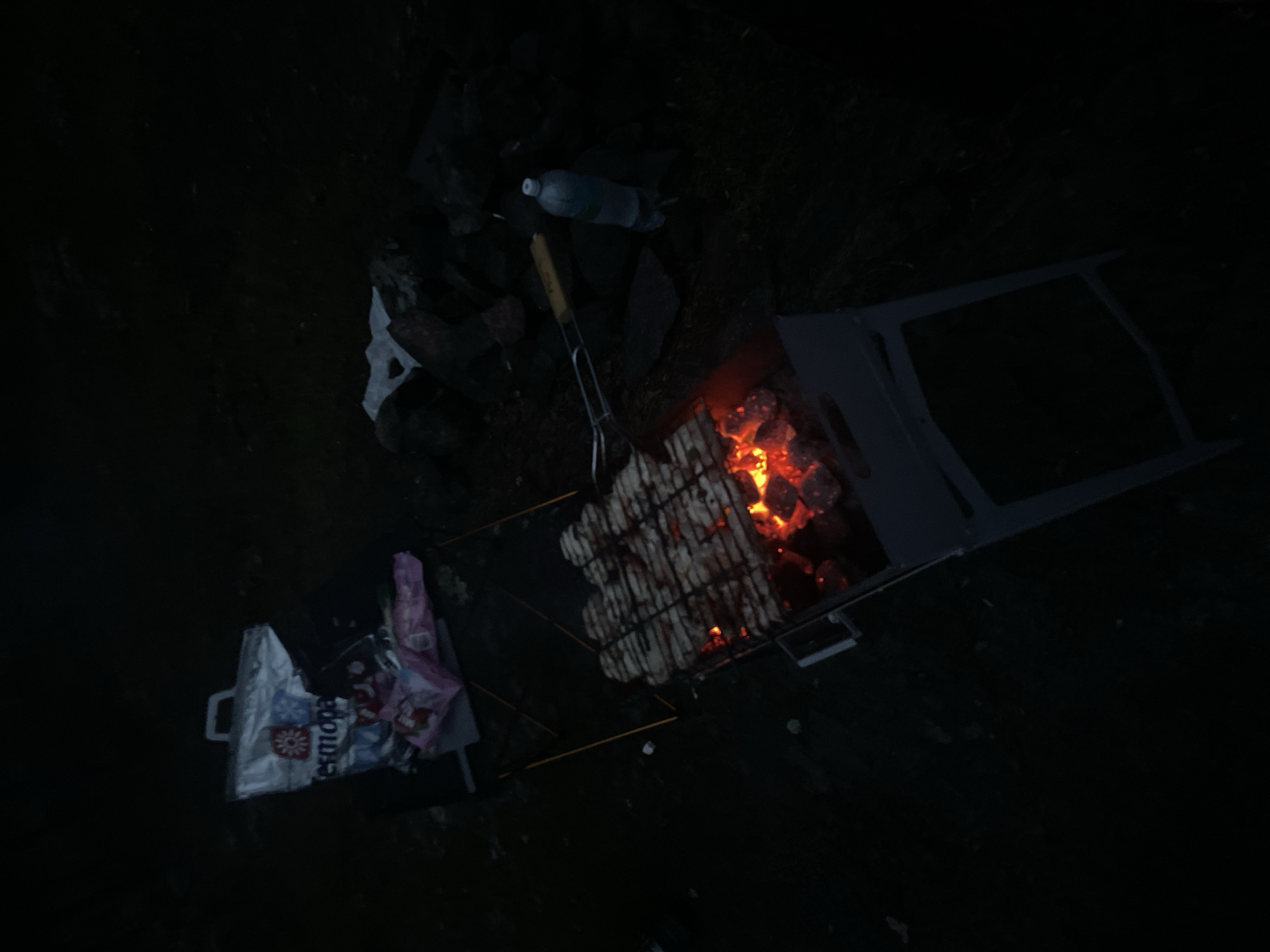 The image captures an outdoor night scene in the city of Yarkoe, Russia. The central focus is a dark gray metal barbecue grill, or mangál, containing actively glowing red charcoal briquettes, serving as the primary light source for the immediate area. Positioned over the hot coals is a metal grate holding numerous pieces of light-colored meat, appearing to be in the early stages of grilling. To the upper left, a silver-colored shopping bag, featuring the prominent white text "Tesmopa" and a red floral design, rests on a dark, flat surface. Adjacent to it is crumpled pink plastic packaging, possibly containing food or snacks. To the right of the grill, a clear plastic water bottle with a light blue cap stands upright on uneven, dark, rocky terrain. Next to the bottle, a barbecue tong with a yellow handle is resting. The surrounding environment is unlit, characterized by dark, uneven ground composed of earth and various rocks, with sparse, dark vegetation visible in some areas. No individuals are present in the scene.