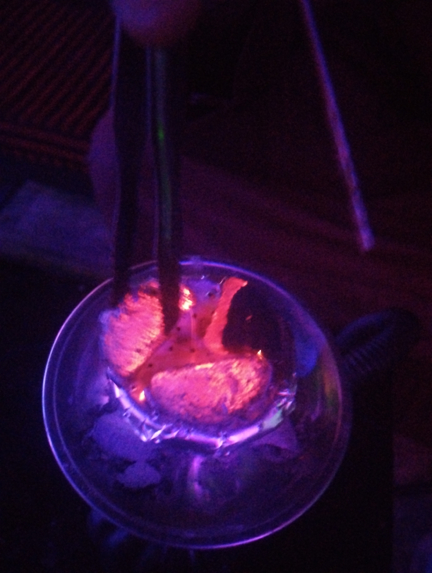 The image shows a hookah with lit coals, glowing with pink and purple light. Two tubes emerge from the hookah, one dark and one green. The background is dark, but a striped surface is visible at the top. There is no indication of the city Tumfure, Nigeria in this picture.