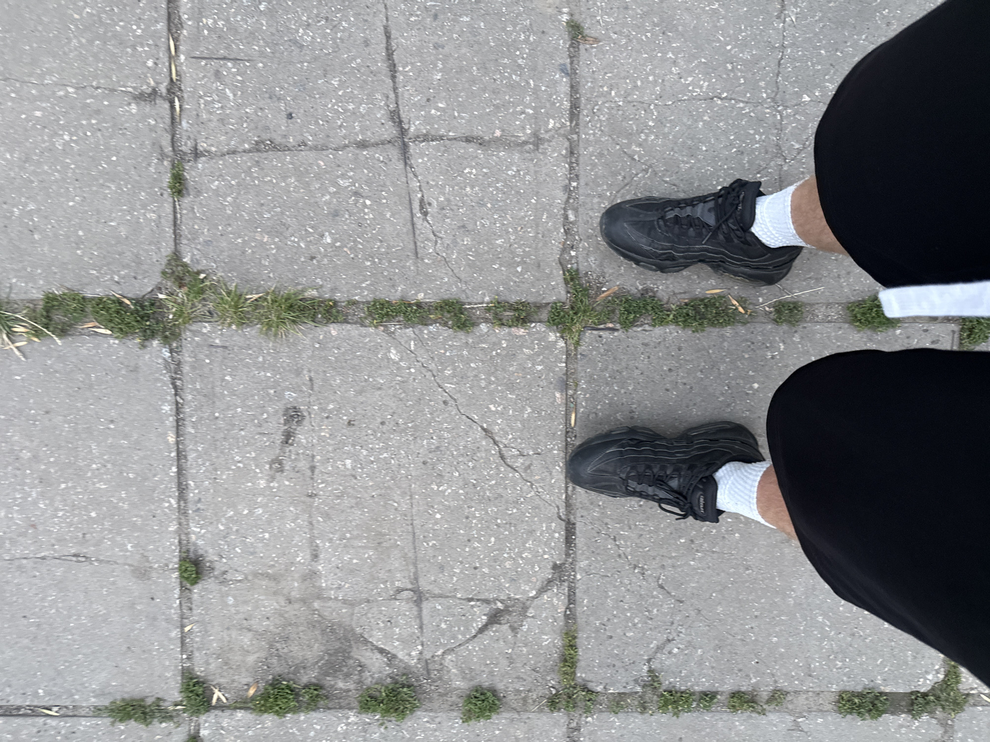 This is a high-angle, medium shot of a person's legs and feet standing on a sidewalk made of gray concrete slabs. The slabs are cracked and uneven, with patches of green grass growing in the cracks. The person is wearing black shorts, white socks, and black athletic shoes. The shoes have thick soles and a textured upper. The lighting suggests it is daytime, possibly overcast. The scene is devoid of any identifying landmarks, vehicles, or other contextual elements that would specifically place it in Zaporizhzhia, Ukraine.