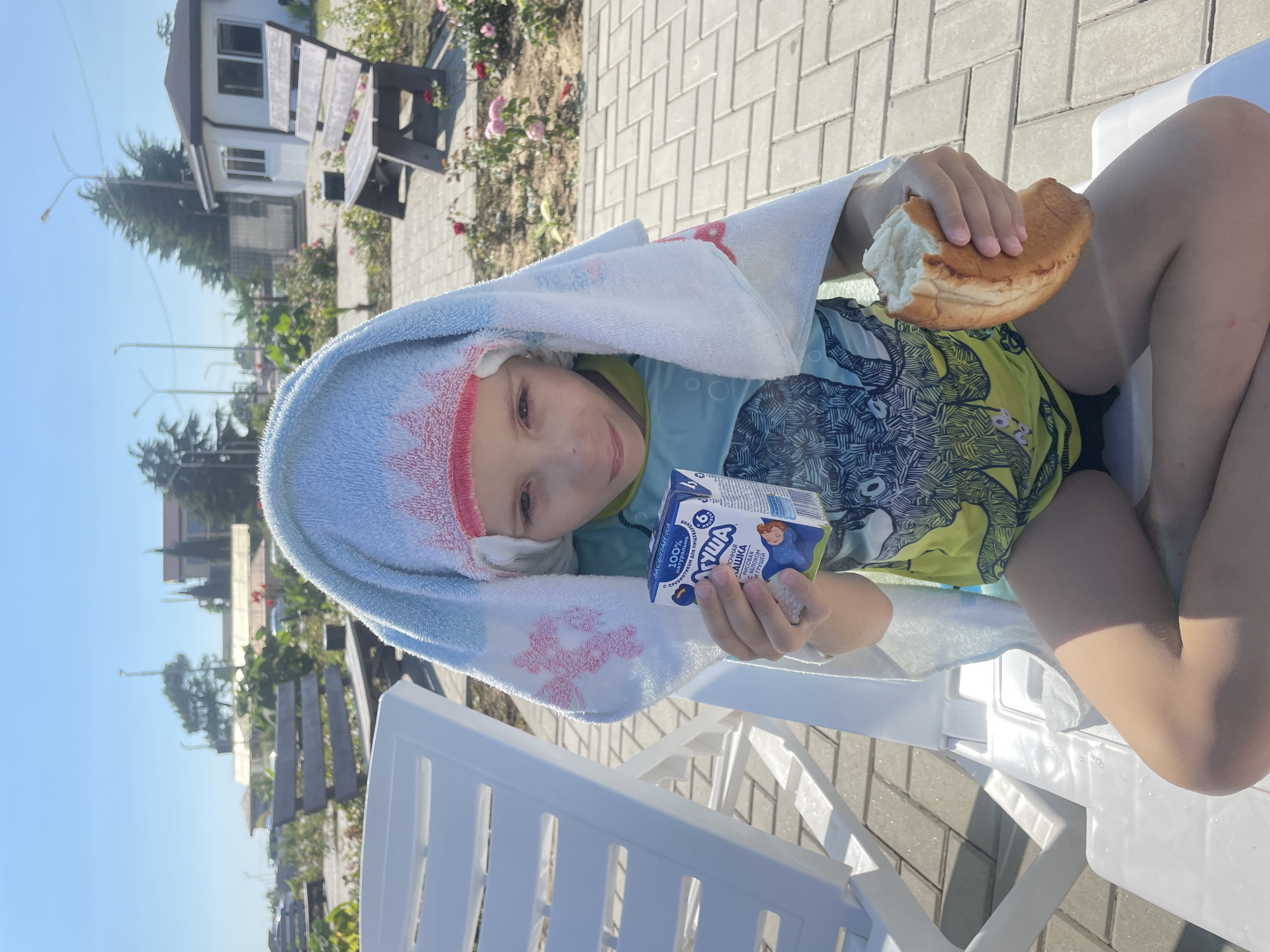 A child is sitting on a white lounge chair outdoors, with a blue and white towel draped over their head. The child is holding a small carton of juice in their left hand and a piece of bread in their right hand. They are wearing a brightly colored shirt with a graphic print. In the background, there are benches, trees, and a building, suggesting a public outdoor area, possibly near a resort or park. The ground is paved with interlocking stones. The lighting indicates a sunny day. The location is identified as Giachrypsh, Abkhazia.