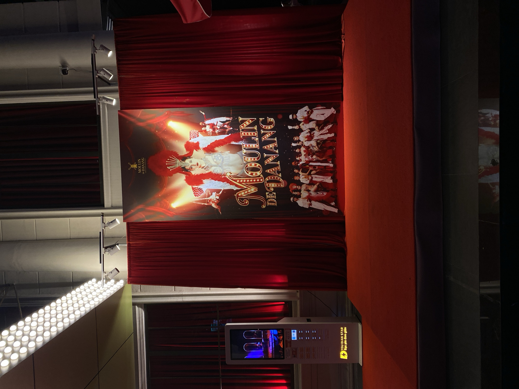 The image shows the exterior of the Moulin Rouge Danang entertainment venue, located in the coastal city of Da Nang, Vietnam. A large red curtain drapes the facade, framing a promotional banner for the show. The banner features a glamorous woman in a feather headdress and a chorus line of performers in elaborate costumes, all set against a fiery orange and yellow background. To the left of the banner, a modern digital kiosk stands on a red carpeted stage, displaying a video of a performance. Spotlights illuminate the scene, casting a dramatic glow on the velvet curtains and the brightly lit banner. The overall impression is one of theatricality and entertainment.