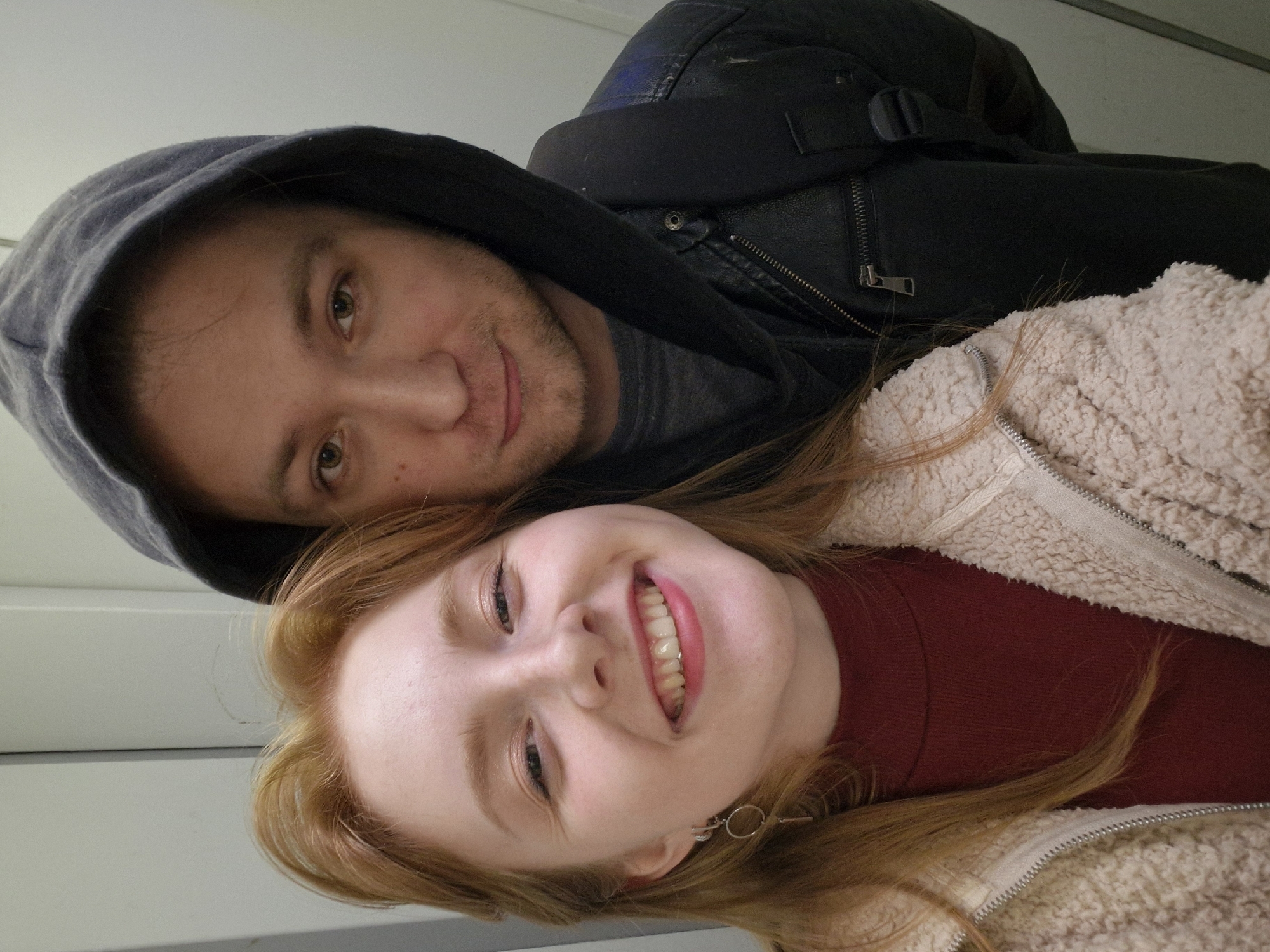 This vertically oriented image captures a close-up selfie of two individuals, a man and a woman, positioned with their heads touching and facing the camera. They appear to be indoors, set against a simple, light-colored background, possibly a wall or the interior of a vehicle.
The woman is in the foreground, on the right side of the frame, with fair skin and light reddish-brown hair. She is smiling widely, showing her teeth, and has a joyful expression. She wears a dark red top beneath a cream or off-white sherpa-style zipped jacket. A small, circular earring with a key-like charm is visible on her right earlobe.
The man is behind and to the left of the woman, with light brown hair and subtle facial stubble. He wears a dark grey hoodie and a black jacket, which appears to be leather, over a grey t-shirt. A strap from a dark backpack or bag is visible over his shoulder. He has a gentle smile, looking content, and his eyes are a light brown or hazel color.
Their close proximity and happy expressions suggest an intimate and affectionate relationship. The lighting is soft and appears to be indoors. There is no visible text in the image. Although the location context of Yekaterinburg, Russia, is provided, the image itself does not offer specific visual cues to identify this particular city. The overall mood of the photograph is happy and warm.