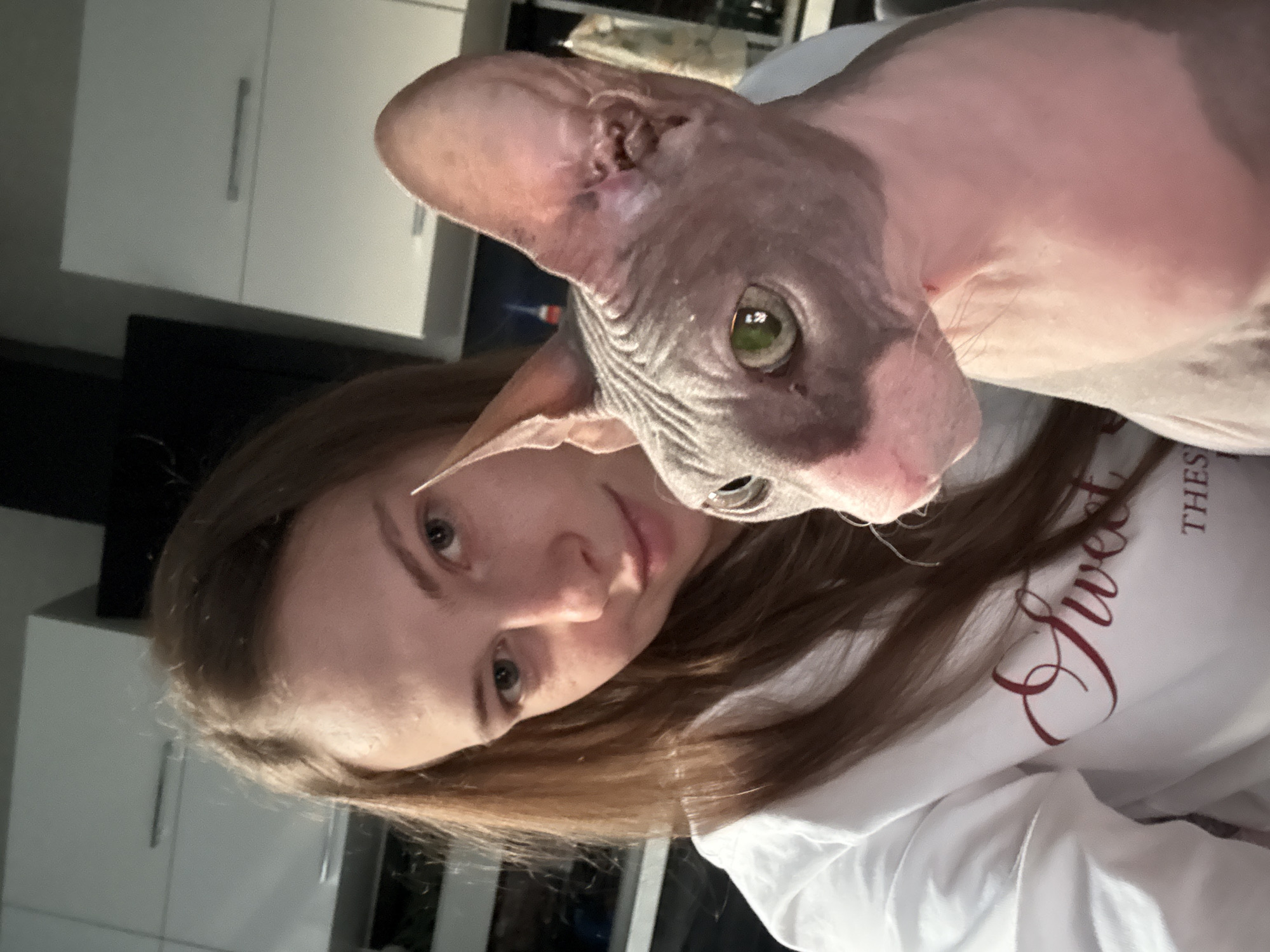 The image captures a close-up, selfie-style shot of a young woman and a Sphynx cat. The woman, with long, light brown hair and light eyes, is wearing a white sweatshirt with dark red lettering that partially reads "Sweet" and "THES". She is looking towards the camera with a soft smile. The Sphynx cat, with its characteristic hairless, wrinkled skin in shades of gray and pink, is positioned to the right of the woman, looking slightly off-camera. Its large ears are prominent. The background appears to be an indoor domestic setting, possibly a kitchen, with white cabinets visible. The lighting suggests an indoor environment, perhaps during the evening or with artificial lighting. No specific details definitively place the image in Novaya Ivanovka, Russia, but it depicts a common domestic scene.
