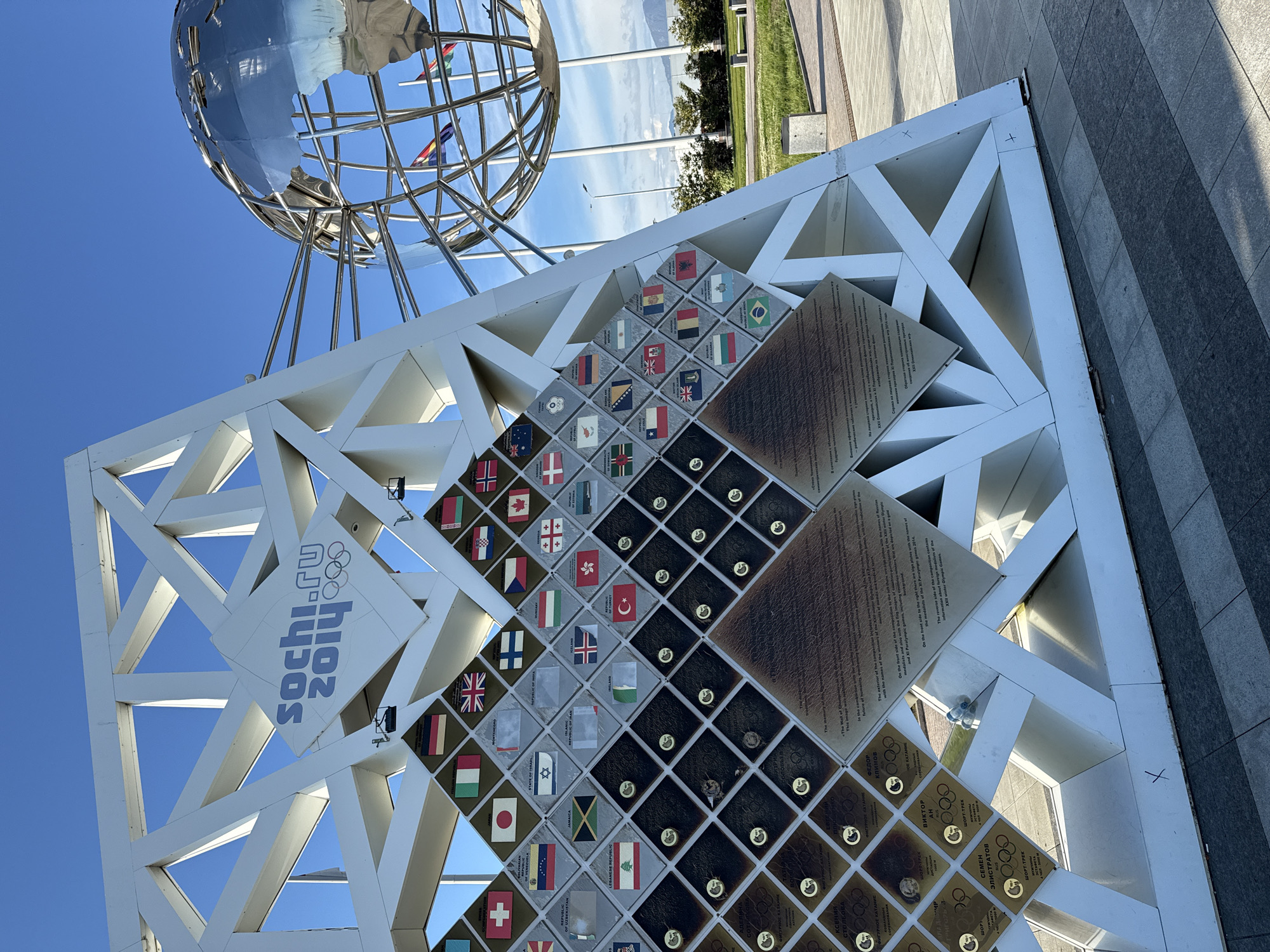 The image depicts a monument dedicated to the Sochi 2014 Winter Olympics and Paralympics in the city of Sirius, Russia. The monument features a silver globe atop a white, geometrically patterned structure. Embedded within the white structure are numerous square tiles displaying the flags of participating nations and commemorative plaques with text. Some plaques display names and depictions related to the Games. The environment is characterized by a blue sky and sections of a paved area, suggesting an outdoor public space.