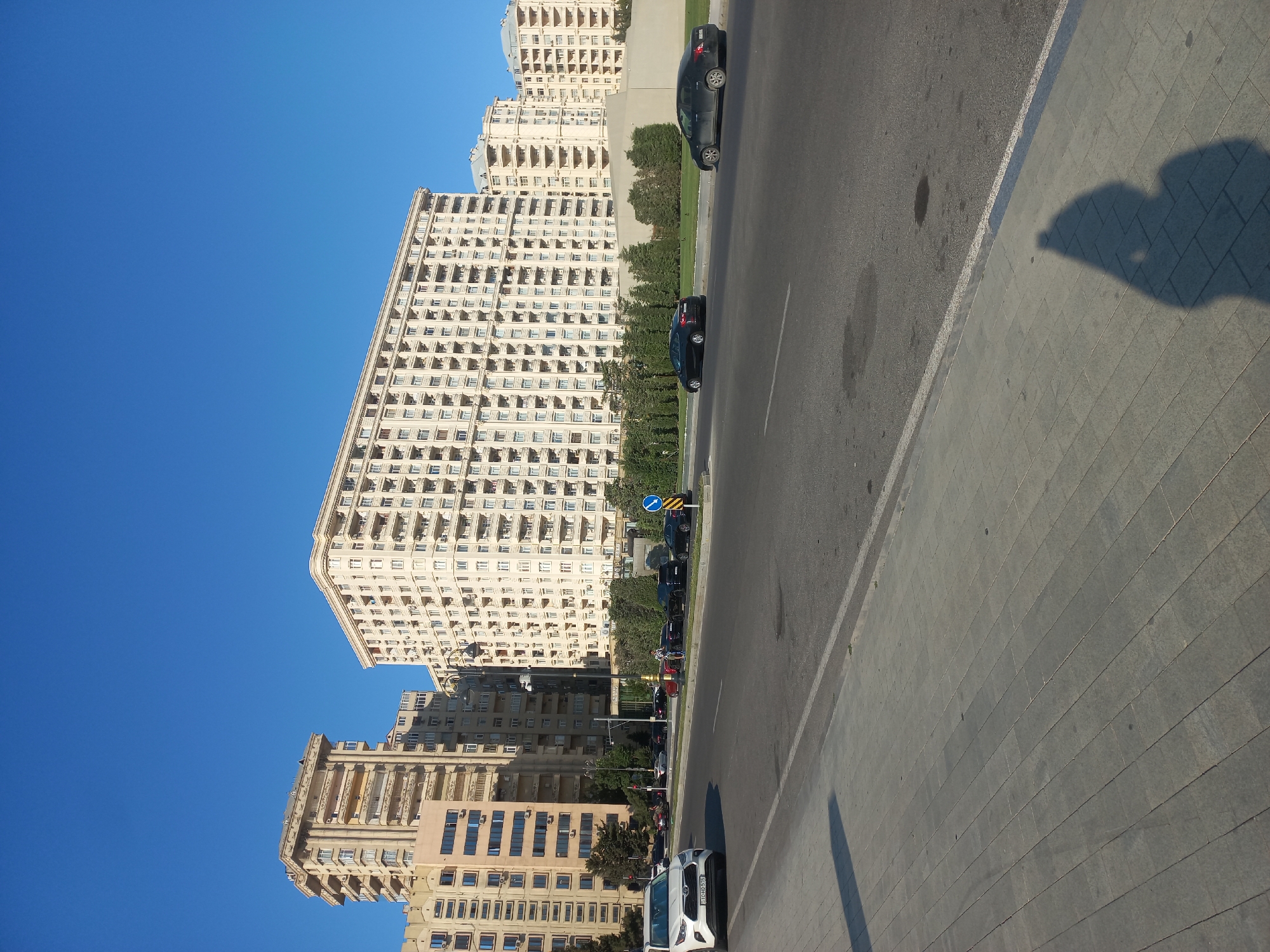 The image depicts a sunny day in Baku, Azerbaijan, with a view of several large, multi-story apartment buildings. The buildings are light-colored and feature numerous balconies and windows, typical of modern residential architecture in the city. In the foreground, a paved sidewalk runs alongside a street with a few cars driving on it. A white SUV is parked on the left side of the street. The sky is a clear, bright blue, indicating good weather. The overall scene suggests a typical urban environment in Baku, characterized by its expansive residential complexes and well-maintained infrastructure.