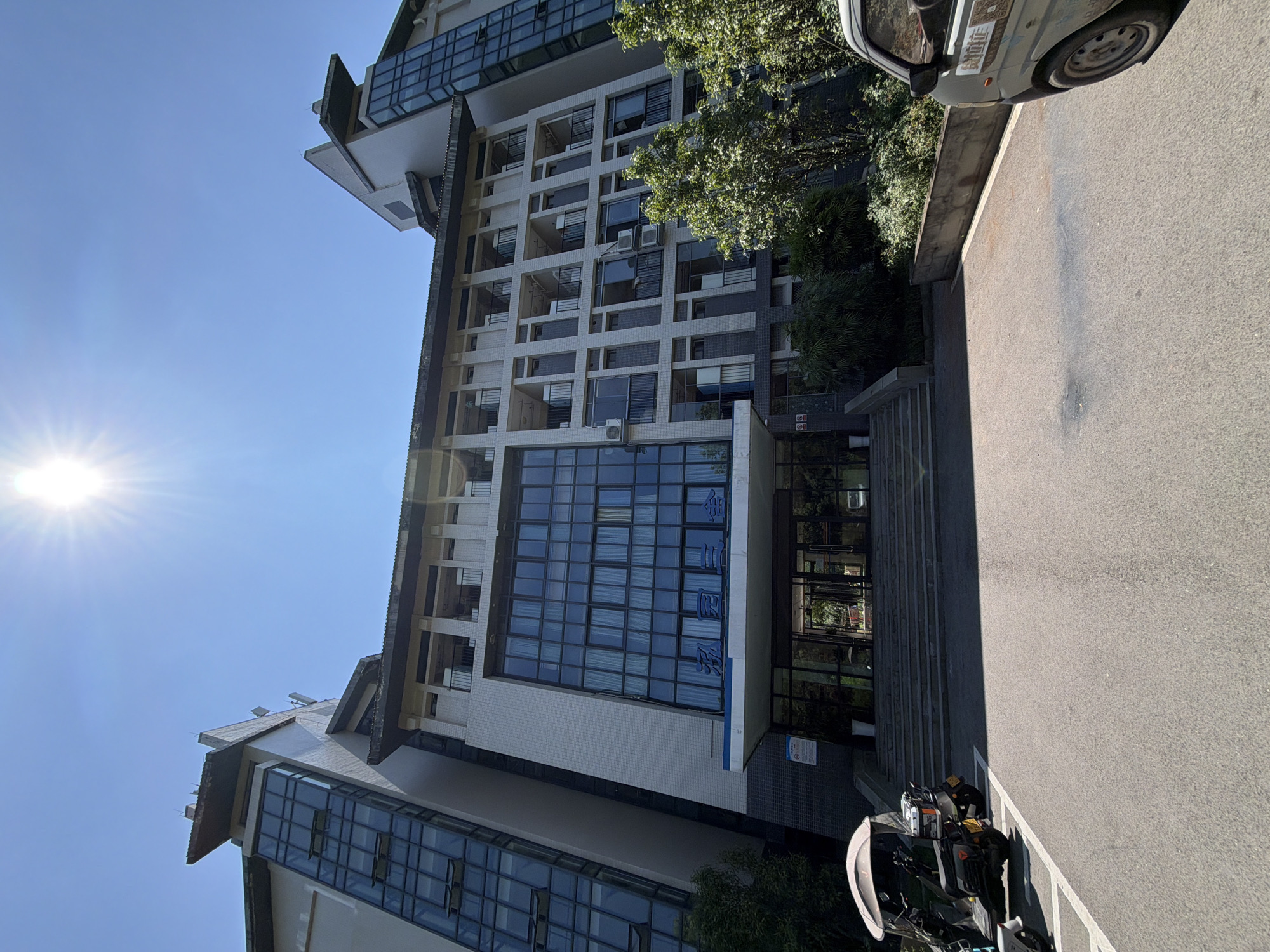 This is a daytime, low-angle exterior shot of a multi-story building, likely an apartment complex or dormitory, located in the city of Jiangjin, China. The building has a modern architectural style, featuring a combination of light-colored facade, dark-framed windows, and glass-paneled sections. The sun is high in the clear blue sky, casting a bright glare.
In the foreground, a paved area leads up to the building's entrance. There are two three-wheeled scooters parked to the left of the entrance, and the rear section of a light-colored van is visible on the right side of the frame. The building entrance is accessed via a wide set of concrete stairs. Above the entrance, a blue sign with Chinese characters is mounted. The building's facade is characterized by repeating rows of balconies or individual rooms with dark railings, interspersed with windows. Some windows are open, and several air conditioning units are visible mounted on the exterior walls. Lush green trees and bushes are planted around the base of the building, with some encroaching onto the paved areas. The overall scene appears to be a typical urban residential or institutional environment.