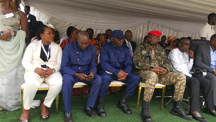The image shows a group of people seated in a semi-circle under a white canopy. The people are seated on gold chairs on a green lawn. The individuals are mostly men and are wearing formal attire, including suits, military uniforms, and traditional clothing. There are a few women in the group wearing elegant dresses. The scene is likely a public event or ceremony, with the attendees seemingly engaged in conversation or listening to a speaker. The location is likely a public park or garden in the city of Rukoko, Democratic Republic of the Congo, based on the provided coordinates (-1.624 latitude, 29.255 longitude).