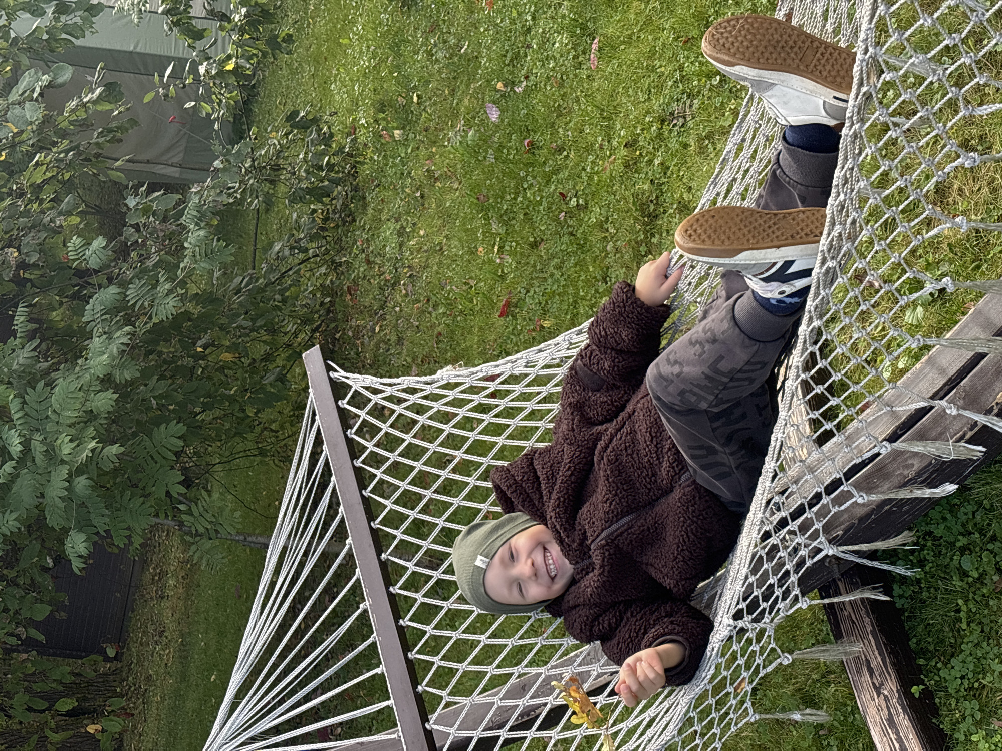 In Рождествено, Russia, a young boy is lying in a hammock outdoors. He is wearing a green beanie, a dark brown fuzzy jacket, and dark pants with a camouflage pattern. He is smiling and holding a yellow leaf in his right hand. The hammock is made of white rope and is suspended between two wooden posts. The background shows a grassy area with fallen leaves and trees. A green tent or structure is visible in the far background behind the trees. The lighting suggests it is daytime.