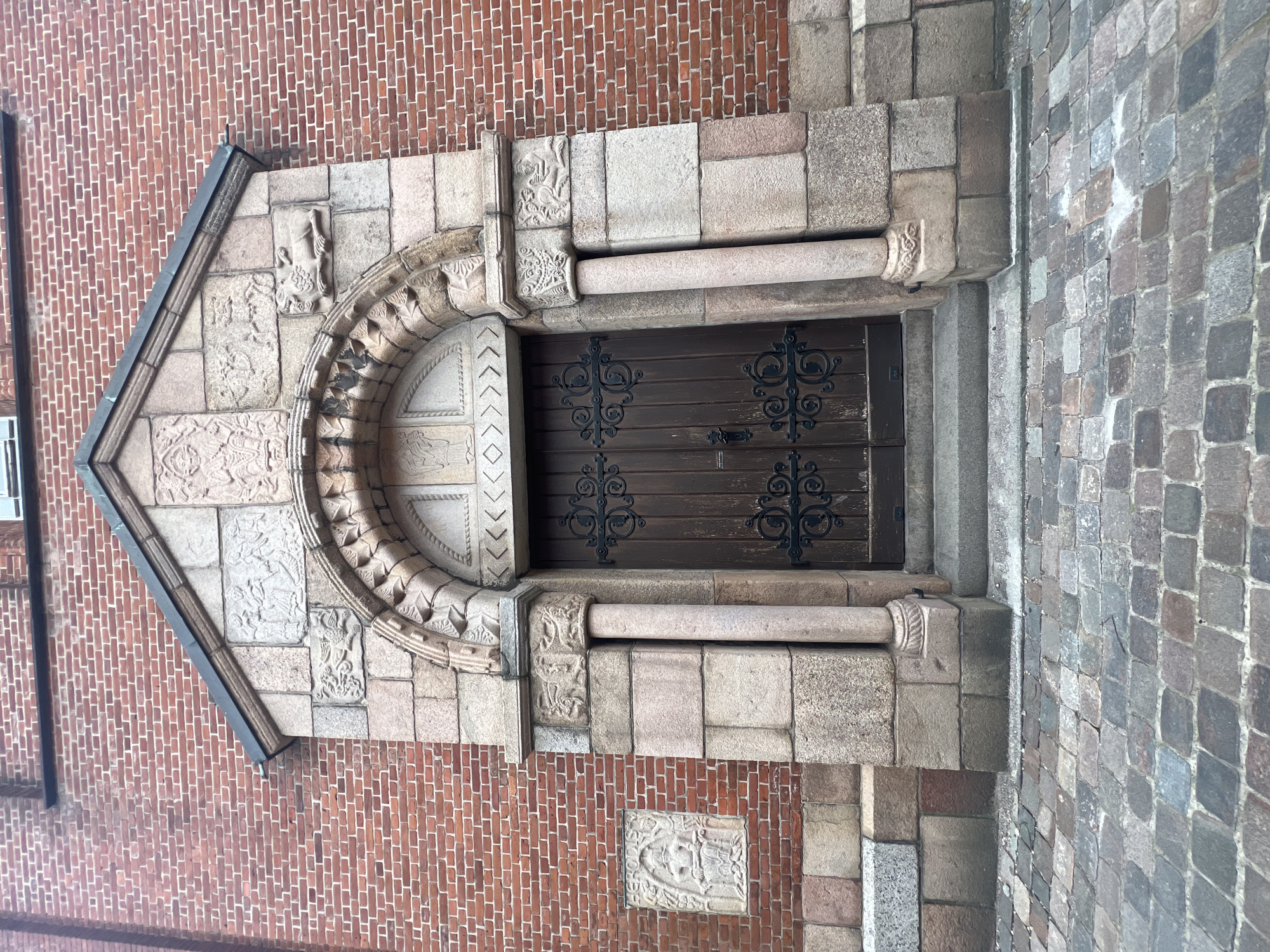 The image shows an exterior view of a building in Aalborg, Denmark. A dark wooden door with black wrought-iron hardware is centrally located. The doorway is framed by light-colored stone, with carved details and a semicircular arch above. The arch and surrounding stonework include various sculpted figures and ornamental patterns. The building is constructed of red brick, with portions of gray stone visible. The ground surface adjacent to the building is paved with small, irregularly shaped stones. The photograph is taken from a slightly low angle, showcasing the architectural details of the structure.