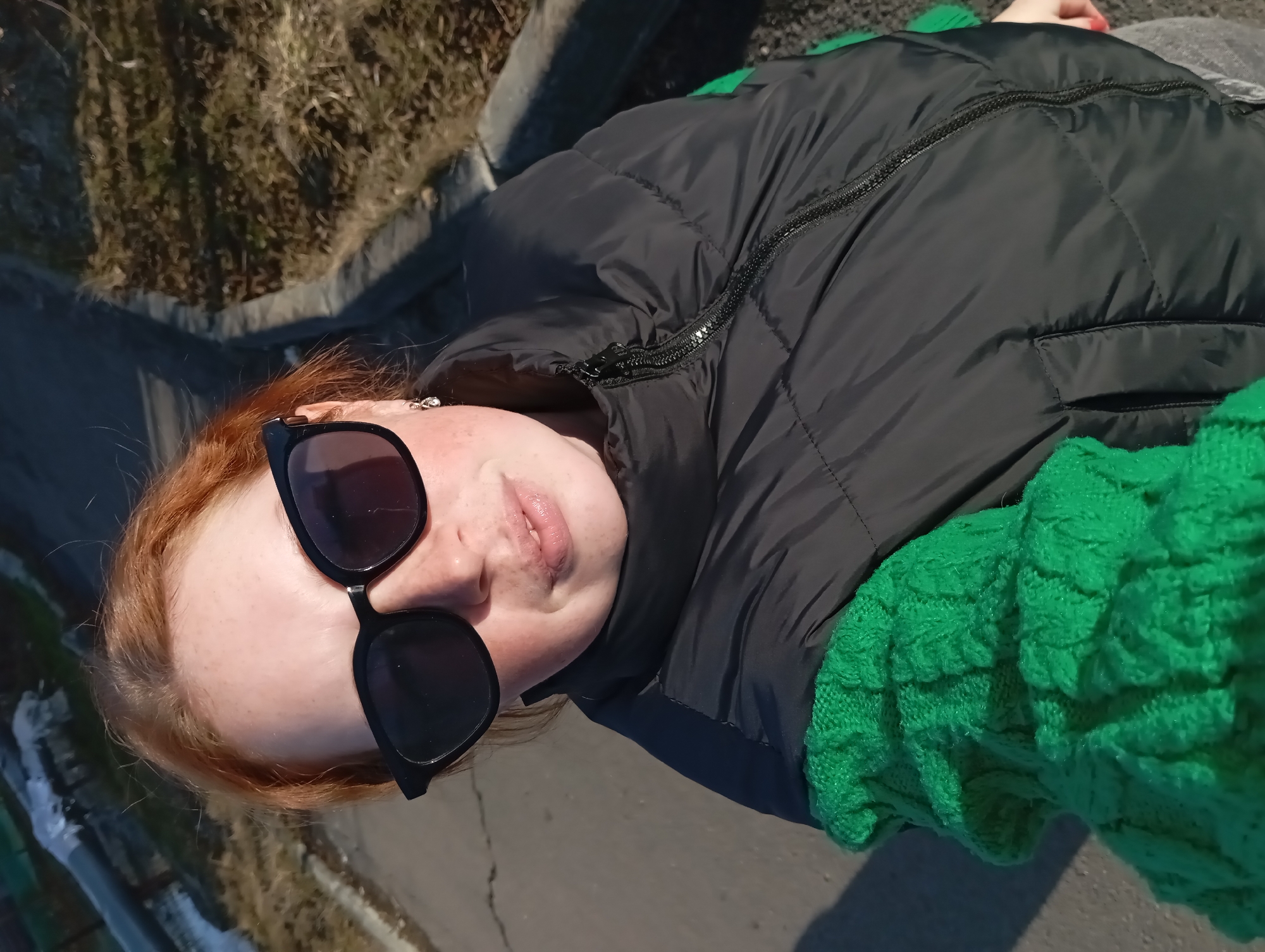 A person with red hair is wearing black sunglasses and a black puffer vest over a bright green sweater. They are outside, standing on a gray paved surface, possibly a sidewalk or road. Patches of dry grass and vegetation are visible along the edge of the pavement. The photo appears to have been taken in Yurgamysh, Russia.