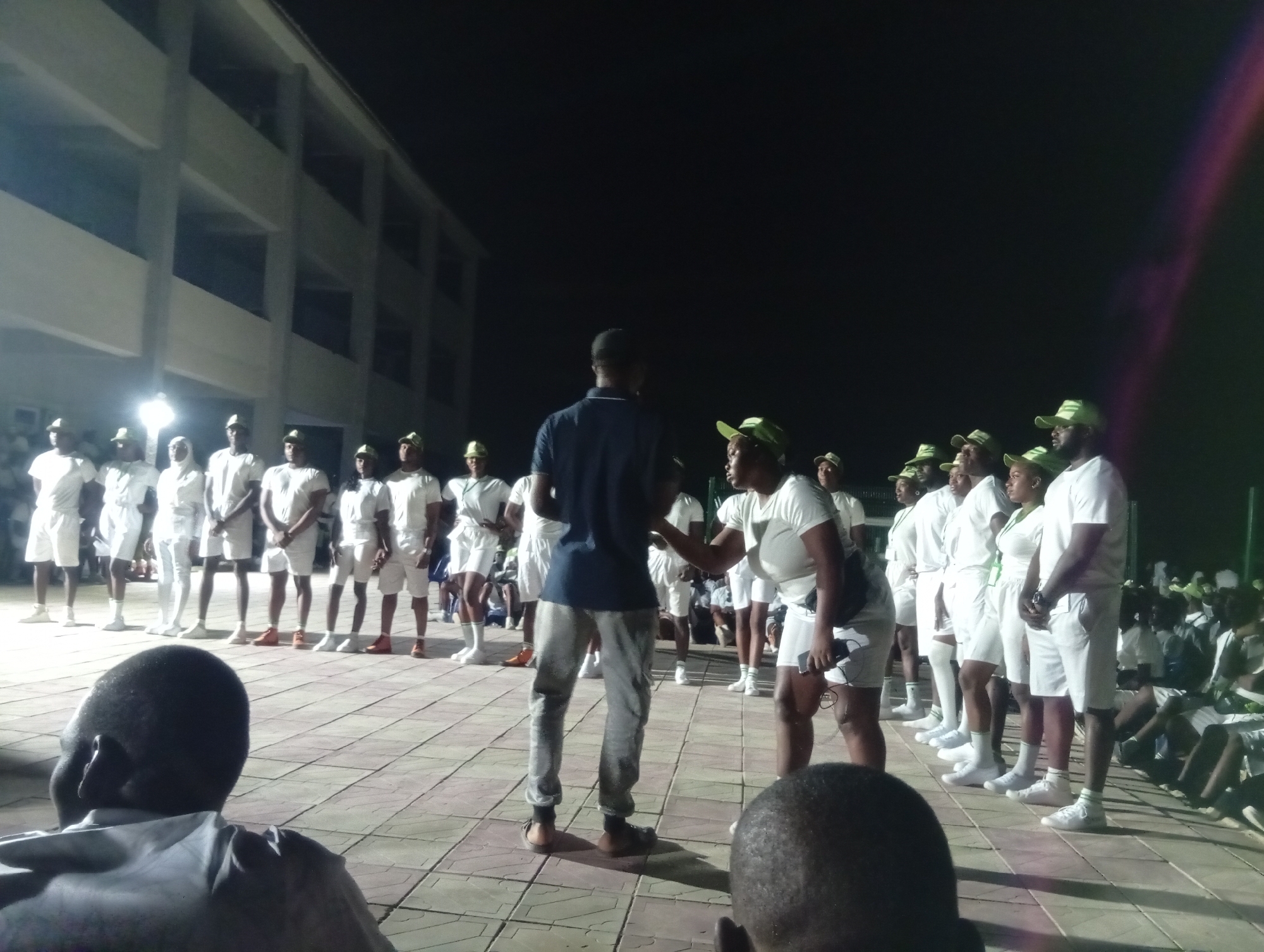 A nighttime outdoor scene shows a group of people gathered on a paved surface in what appears to be Maiduguri, Nigeria. A building is visible in the background. A group of people wearing white shirts and shorts, along with green caps, are standing in a line. In the foreground, two people are interacting, while others are seated and watching. The setting appears to be formal, possibly a ceremony or gathering.