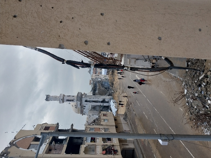 An elevated view captures a street scene in Khan Yunis, Palestinian Territory, under an overcast sky. The foreground on the right features a beige building wall exhibiting multiple bullet holes and vertical utility cables. To the left, a grey lamppost stands beside a pile of concrete rubble and debris. The central midground depicts a wet, asphalt street with faint white markings, suggesting recent precipitation. Several civilians, individually and in small groups, walk along the street, moving away from the viewer.
Flanking the street, buildings show extensive damage. On the left, a multi-story structure has significant structural damage, with its facade partially collapsed, exposing interior rooms and fractured walls. In the background, a prominent mosque with a white minaret and dome is visible, also heavily damaged but structurally upright. The minaret shows visible damage, and surrounding buildings in the background are similarly destroyed or severely compromised.